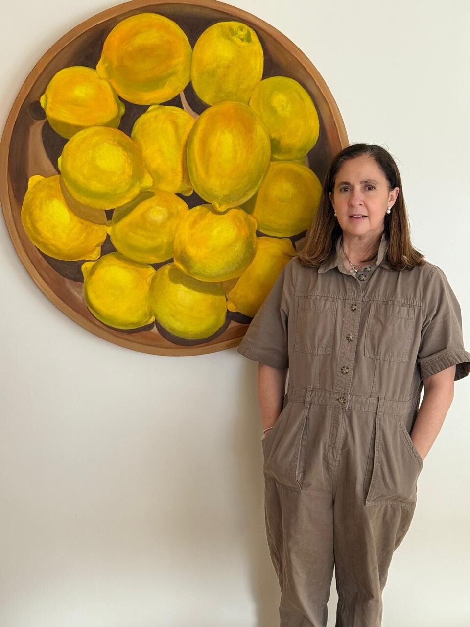 A woman standing in front of a circular painting of lemons on a white wall. She has shoulder-length brown hair and is wearing a tan jumpsuit with her hands in her pockets.
