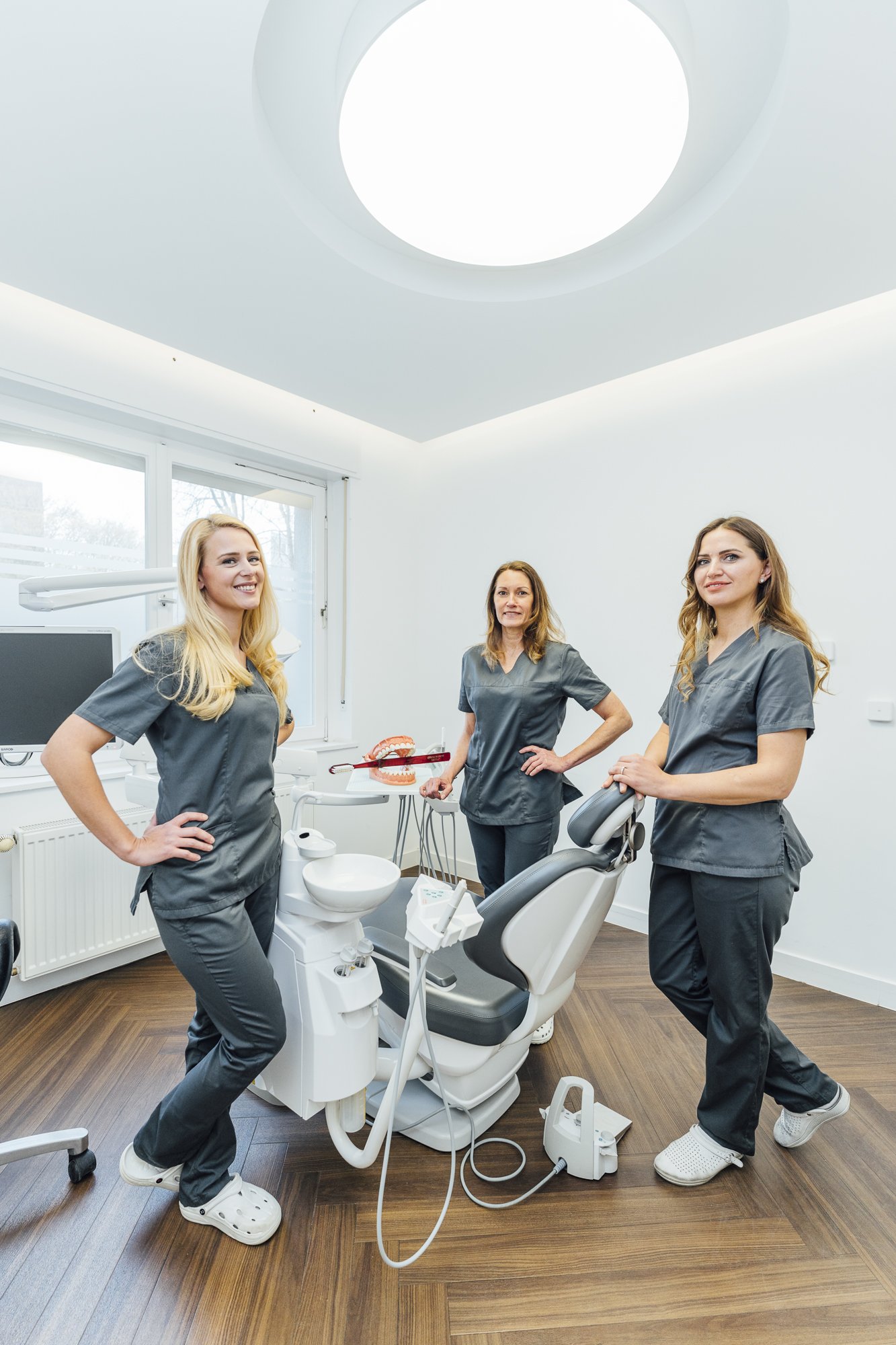 Drei Zahnärztinnen in einer Zahnarztpraxis, die vor einem Behandlungsstuhl stehen, mit zahnmedizinischen Werkzeugen und Modellen in einer hellen, modernen Behandlungslounge.