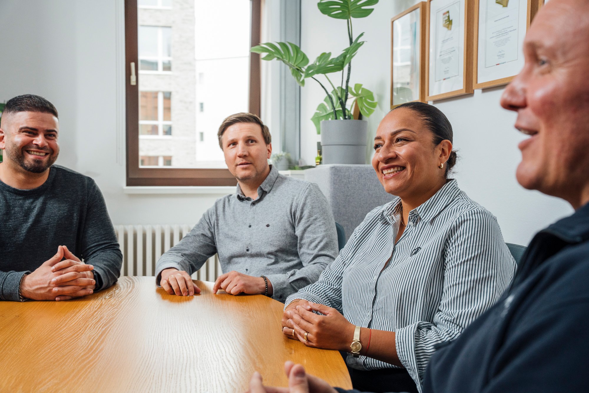 Vier Menschen sitzen an einem Konferenztisch und unterhalten sich in einem modernen Büro unter einem Fenster mit Pflanzen und gerahmten Dokumenten an der Wand.