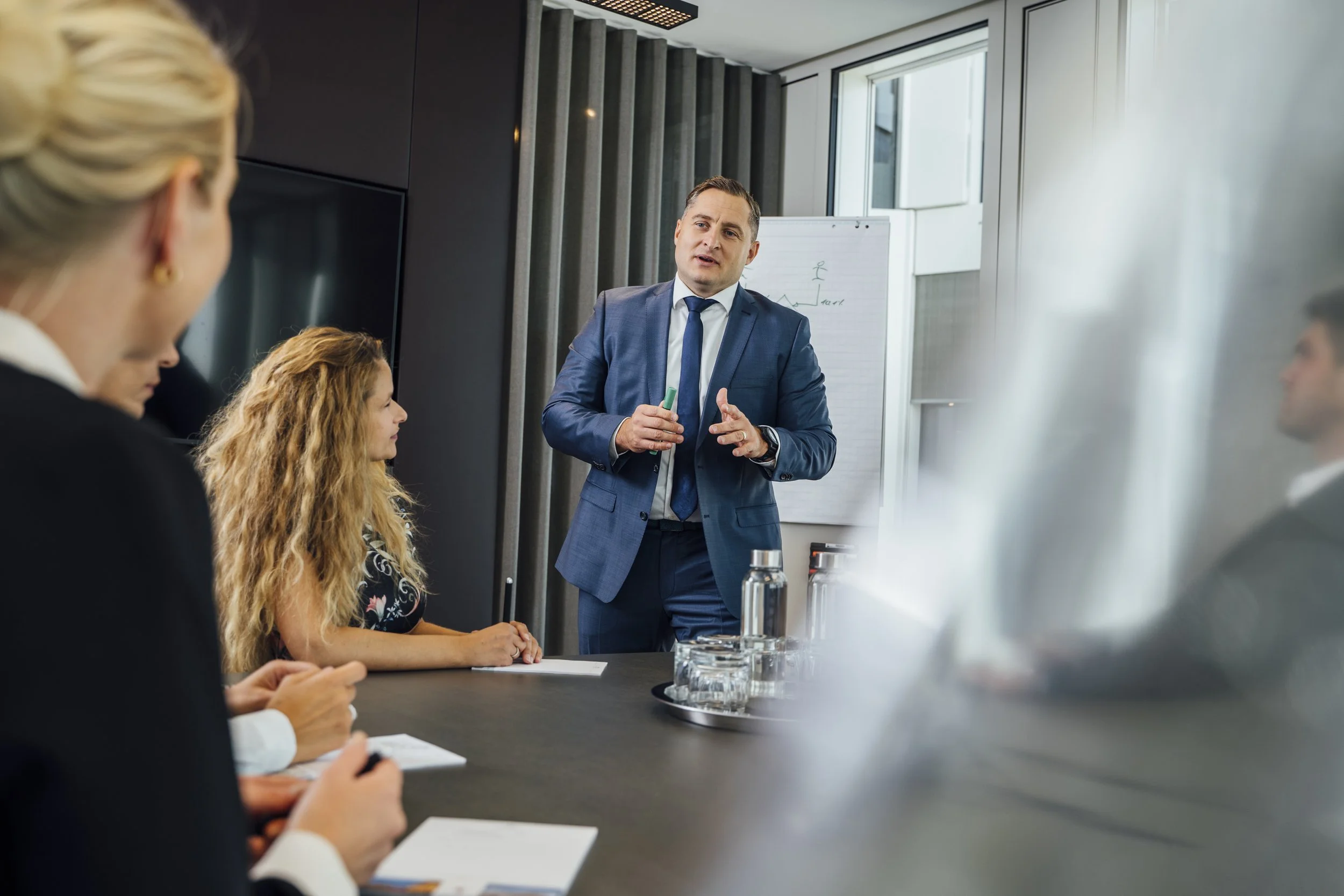 Mann in Anzug erklärt einer Gruppe von Personen in einem Meetingraum.