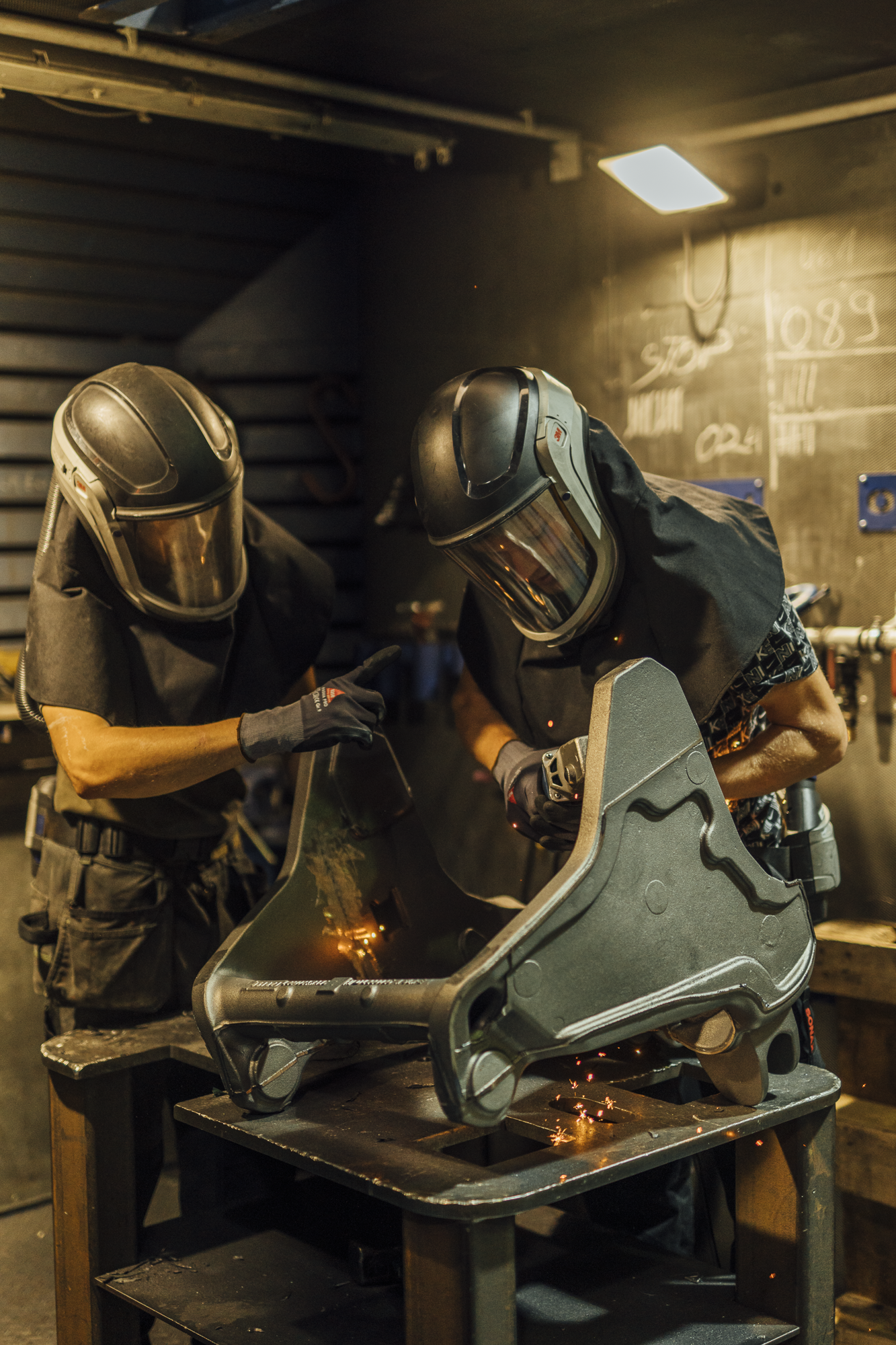 Zwei Personen beim Schweißen eines Metallteils in einer Werkstatt, beide tragen Schutzhelme und schwarze Arbeitskleidung.