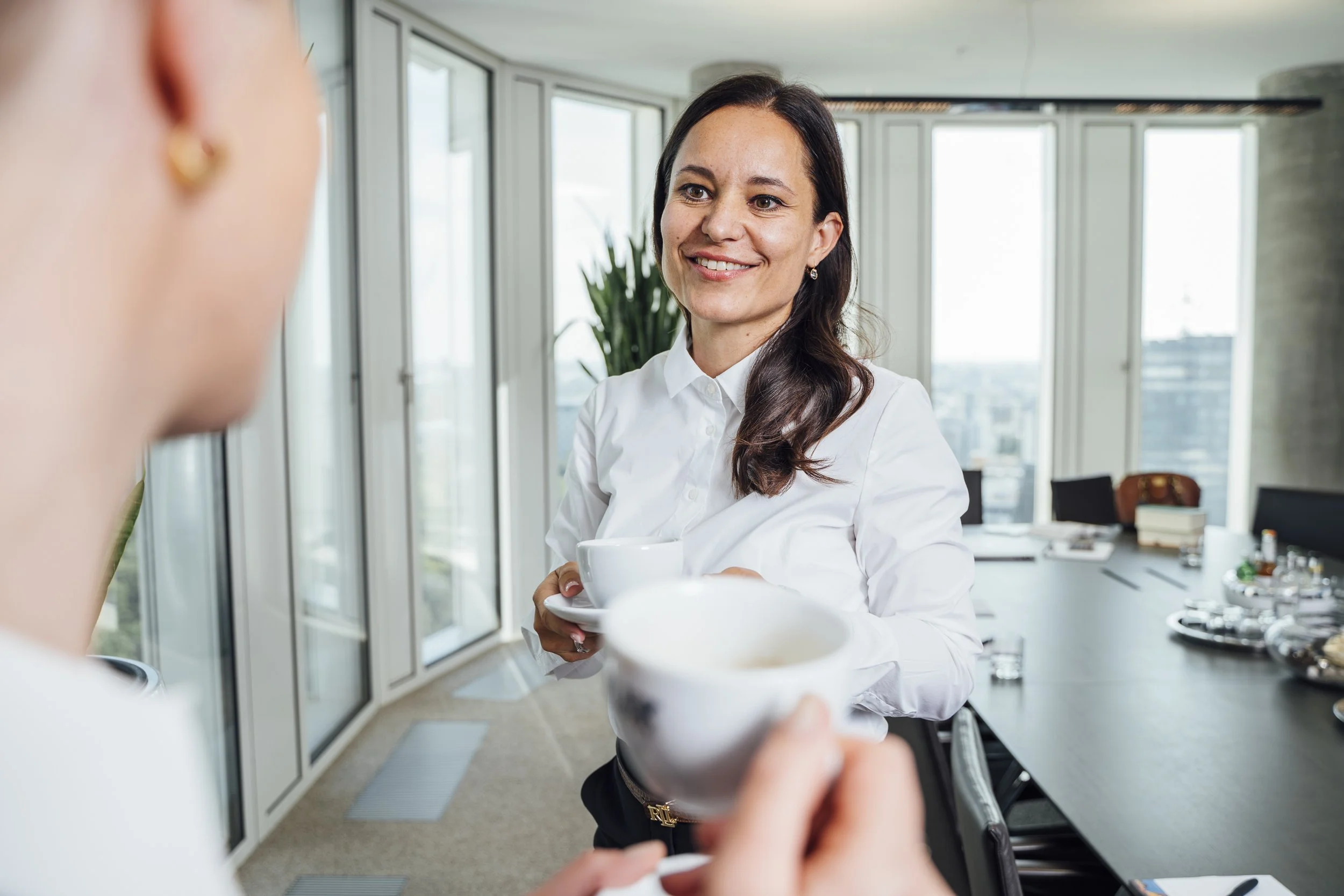 Zwei Frauen tauschen sich in einem modernen Büro mit großen Fenstern aus, eine hält eine Tasse Kaffee und die andere ein Dessert.