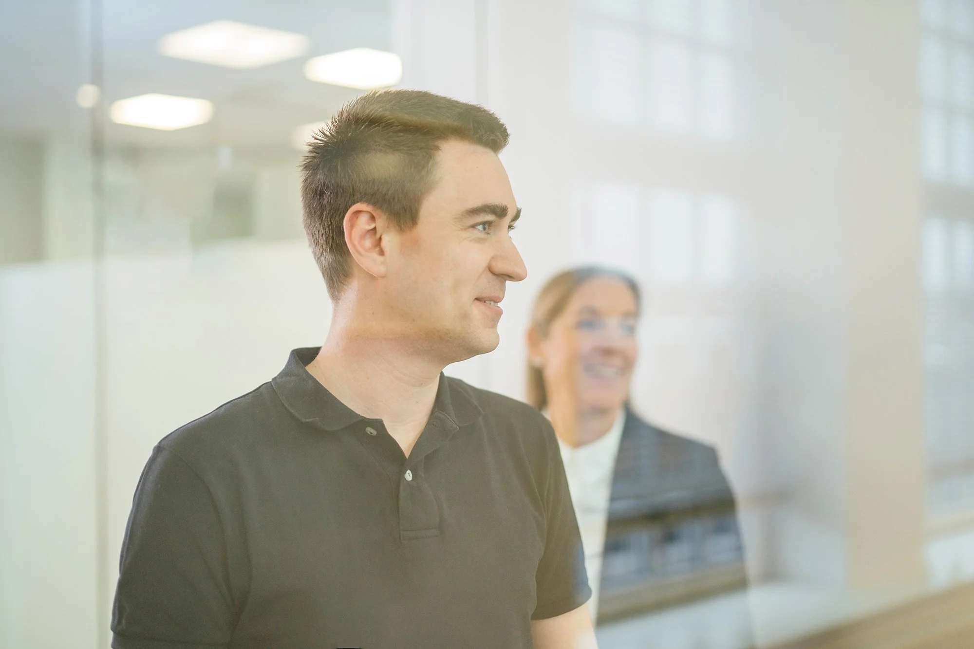 Ein junger Mann mit dunklem Kurzhaarschnitt, trägt ein schwarzes Polo-Shirt, steht vor einer Glaswand, im Hintergrund ist eine lachende Frau zu sehen.