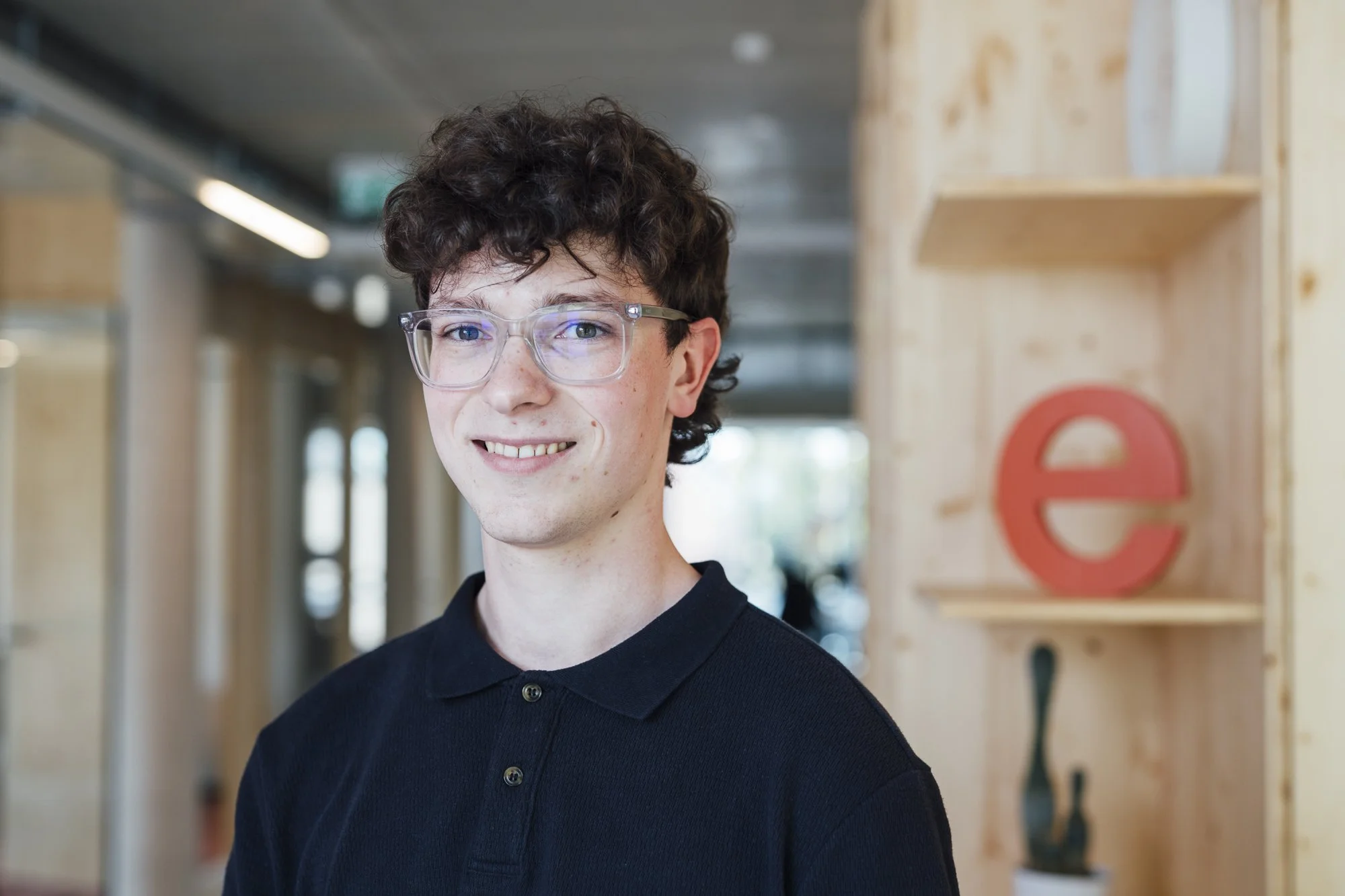 Junge Mann trägt schwarze Poloshirt und klare Brille, steht vor Holzwand mit rotem 'e'-Zeichen in modernem Büro.