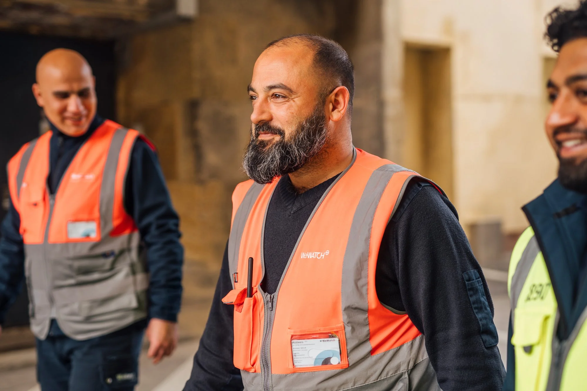 Drei Männer in hoher Sichtschutzkleidung und Sicherheitswesten, lächeln in einem Gebäude mit Backsteinwänden.