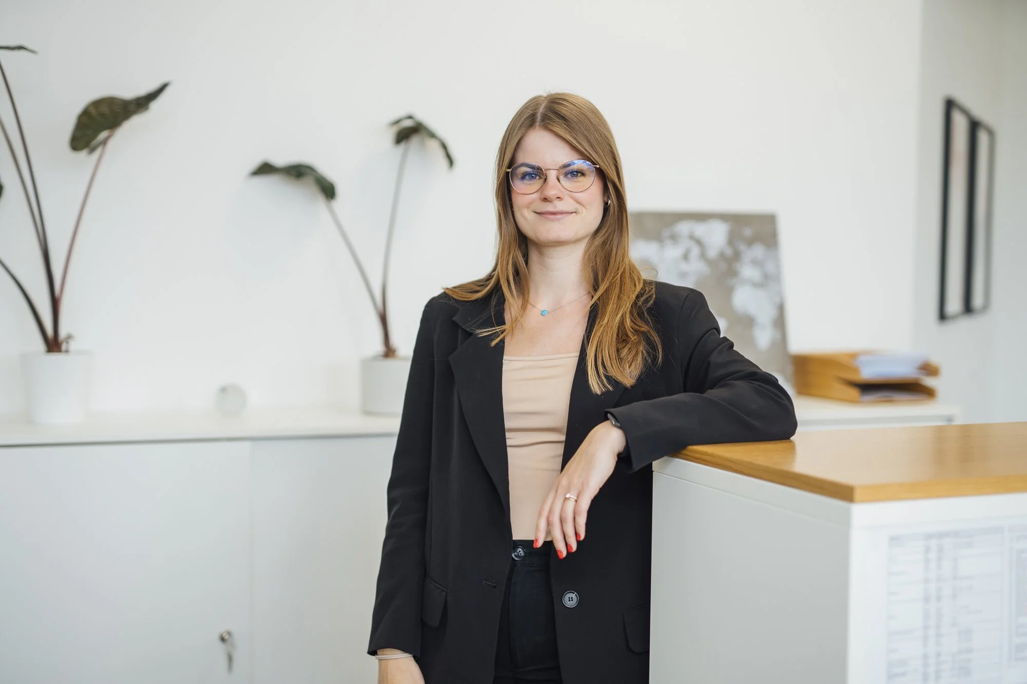 Eine junge Frau mit Brille, schwarzer Jacke und beige Top steht an einem Empfangstresen in einem modernen Büro.