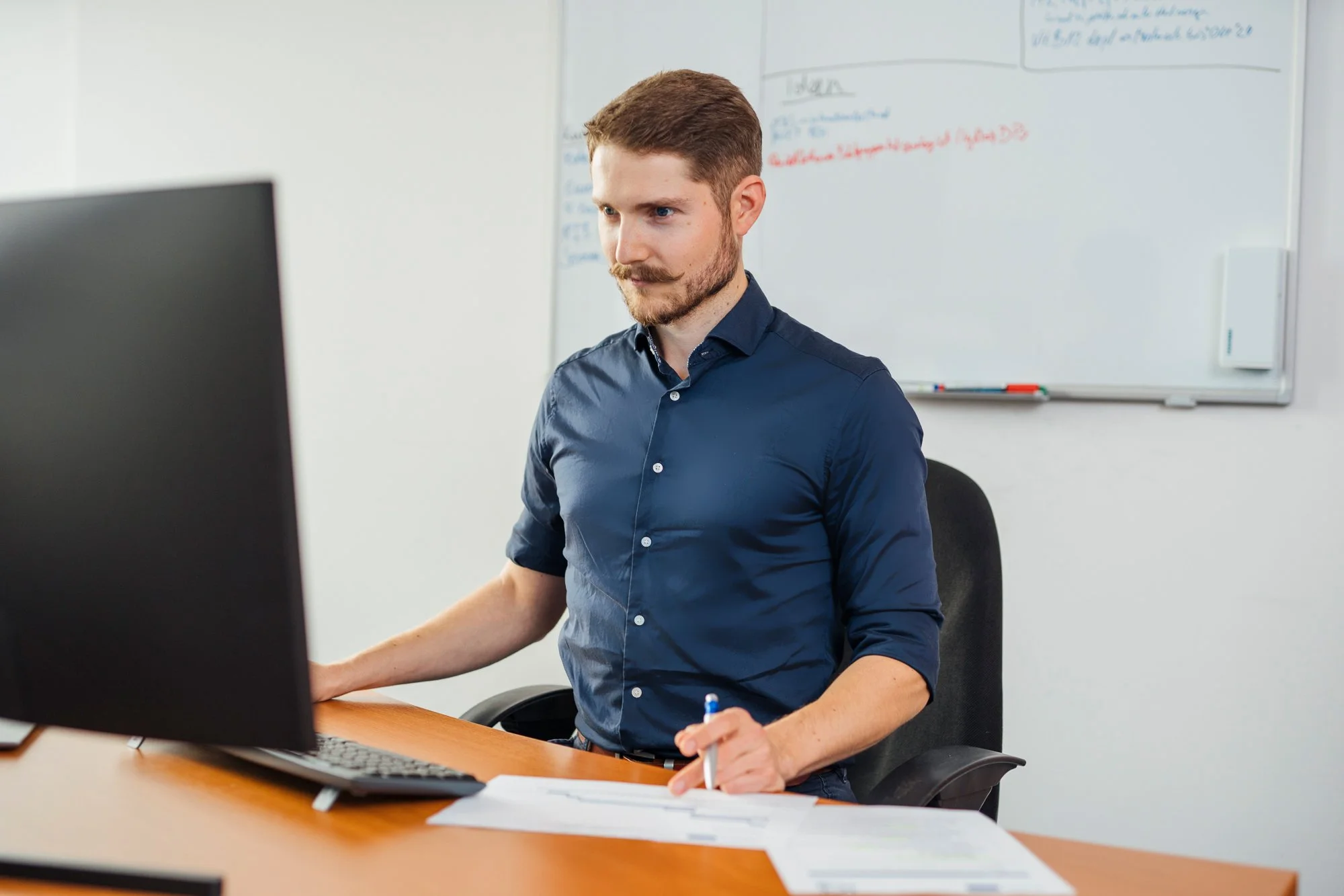 Mann sitzt am Schreibtisch vor einem Computer und schreibt auf Papier, im Hintergrund eine Whiteboard mit Notizen.