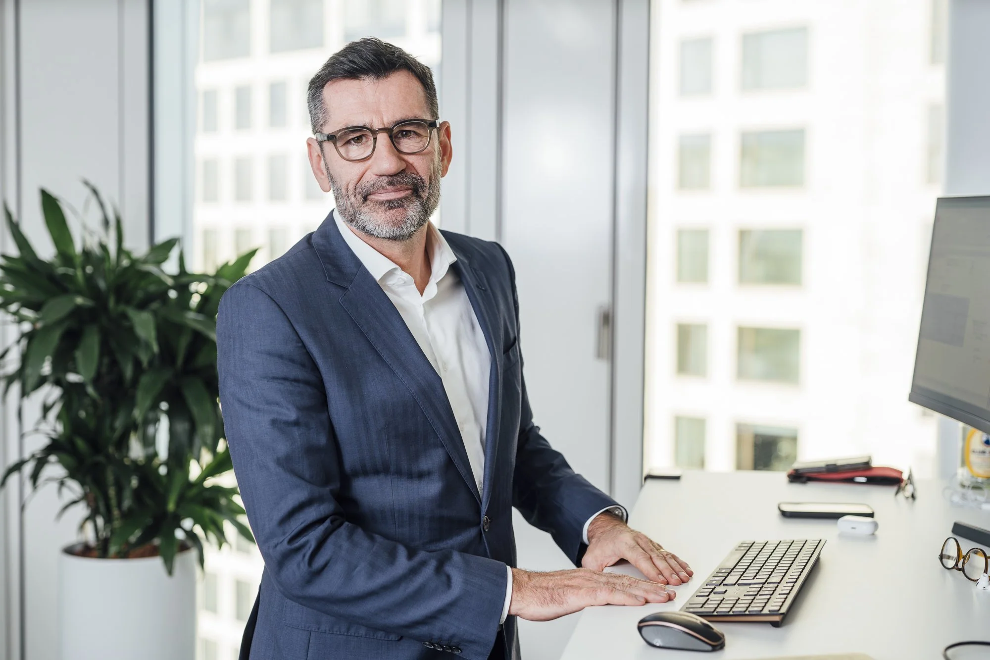 Ein älterer Mann mit Brille und grauem Bart trägt einen dunklen Anzug und sitzt an einem Schreibtisch im Büro, vor einem Bildschirm, mit Blick auf die Stadt durch die Fenster im Hintergrund.