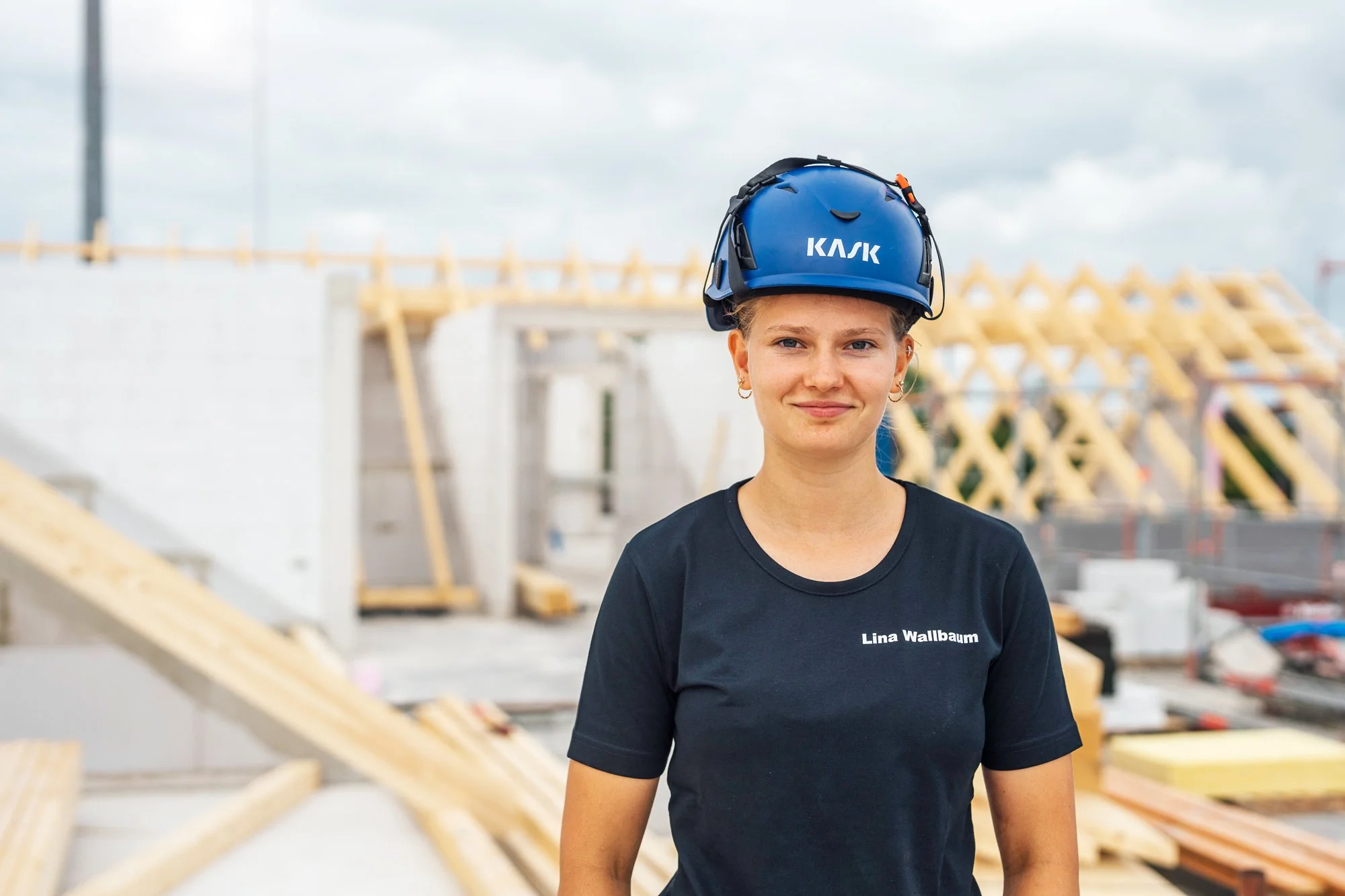 Junge Frau mit Bauhelm auf einer Baustelle, im Hintergrund Holzstrukturen und Baustoffe.