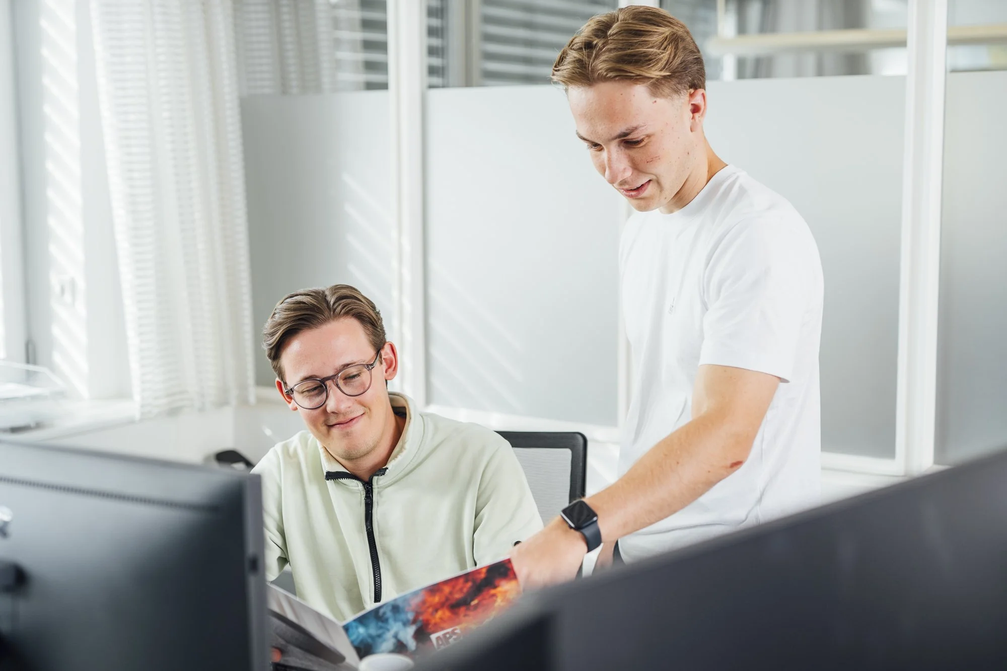 Zwei junge Männer arbeiten gemeinsam an einem Computer in einem modernen Büro, einer der Männer zeigt auf den Bildschirm, beide wirken engagiert und freundlich.