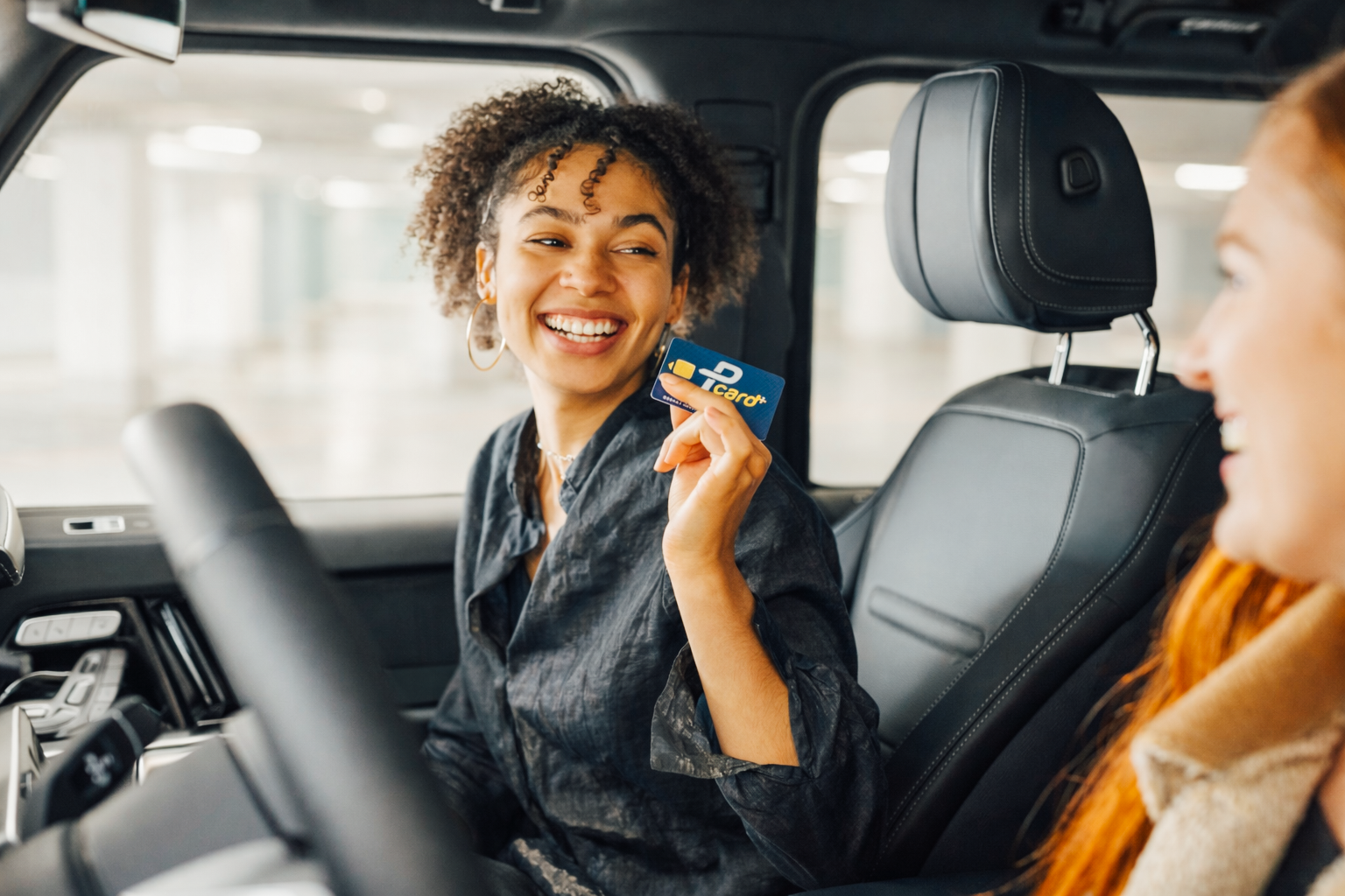 Zwei Frauen sitzen im Auto, die eine hält eine Kreditkarte hoch und lächelt, während die andere ebenfalls lächelt.