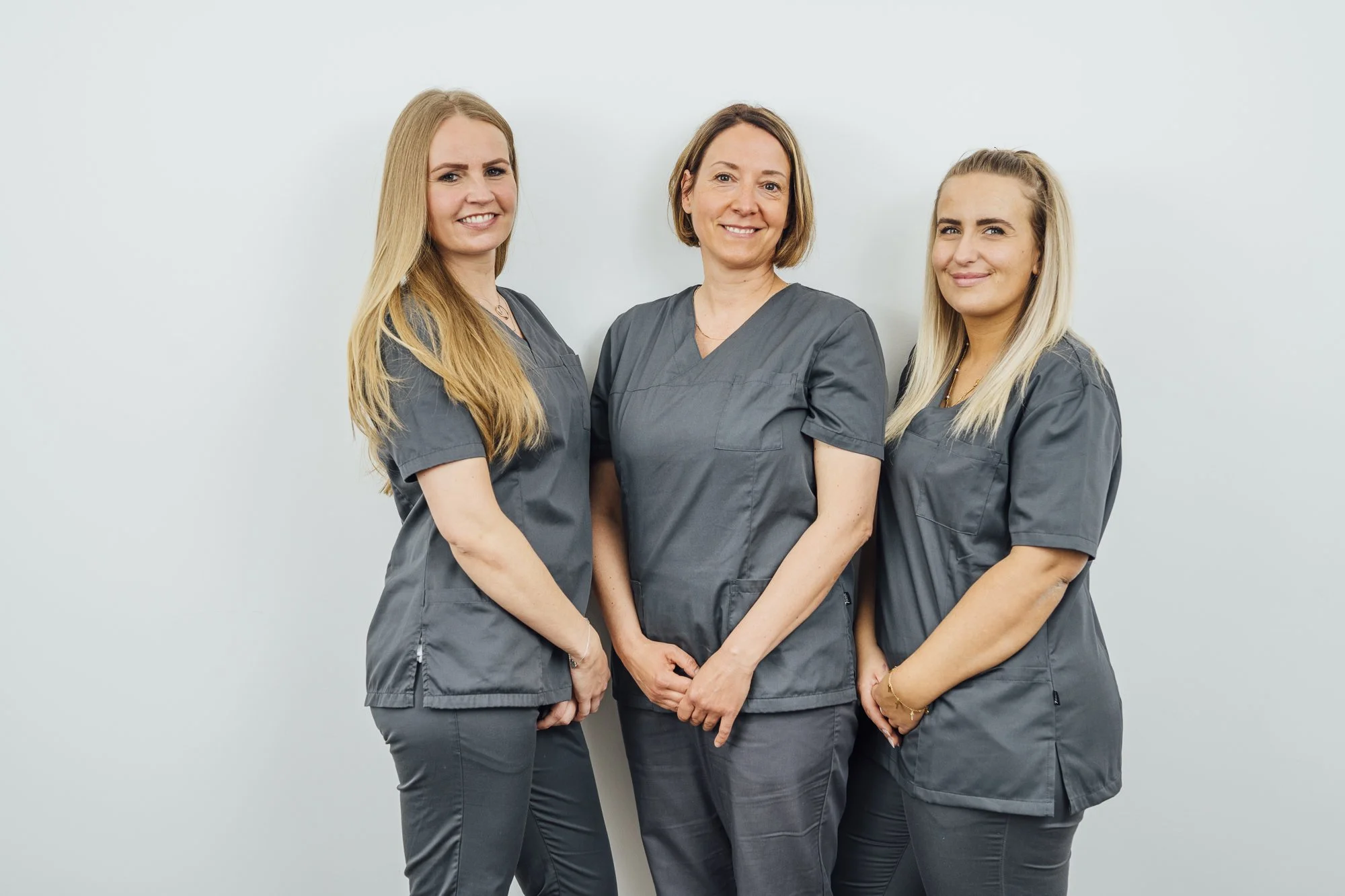 Drei weibliche medizinische Fachkräfte in grauen Uniformen stehen nebeneinander vor einer weißen Wand und lächeln in die Kamera.