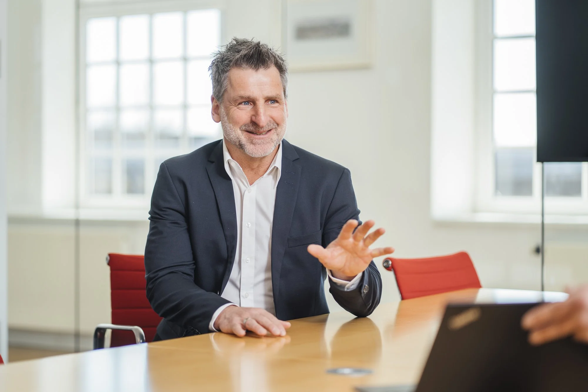 Ein lächelnder, älterer Mann in einem schwarzen Anzug sitzt an einem Konferenztisch, spricht und gestikuliert mit seiner rechten Hand, während er mit jemand anderem spricht.