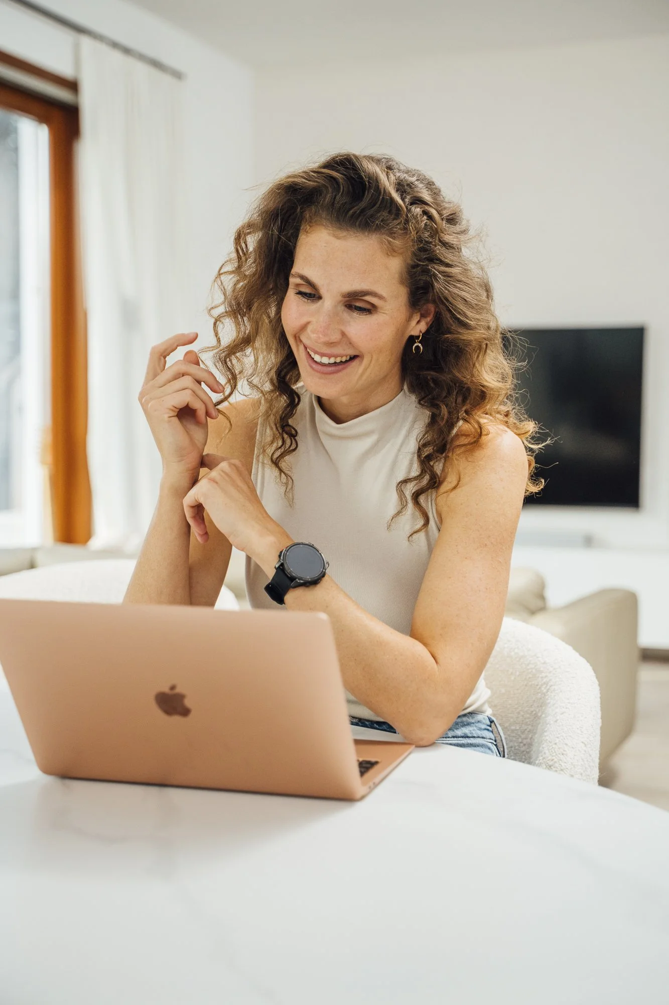Lächelnde Frau mit Locken schaut auf eine roségoldene Apple-Laptop, trägt ein weißes Oberteil, eine Uhr und kleine Ohrringe, sitzt in einem hellen Wohnzimmer.