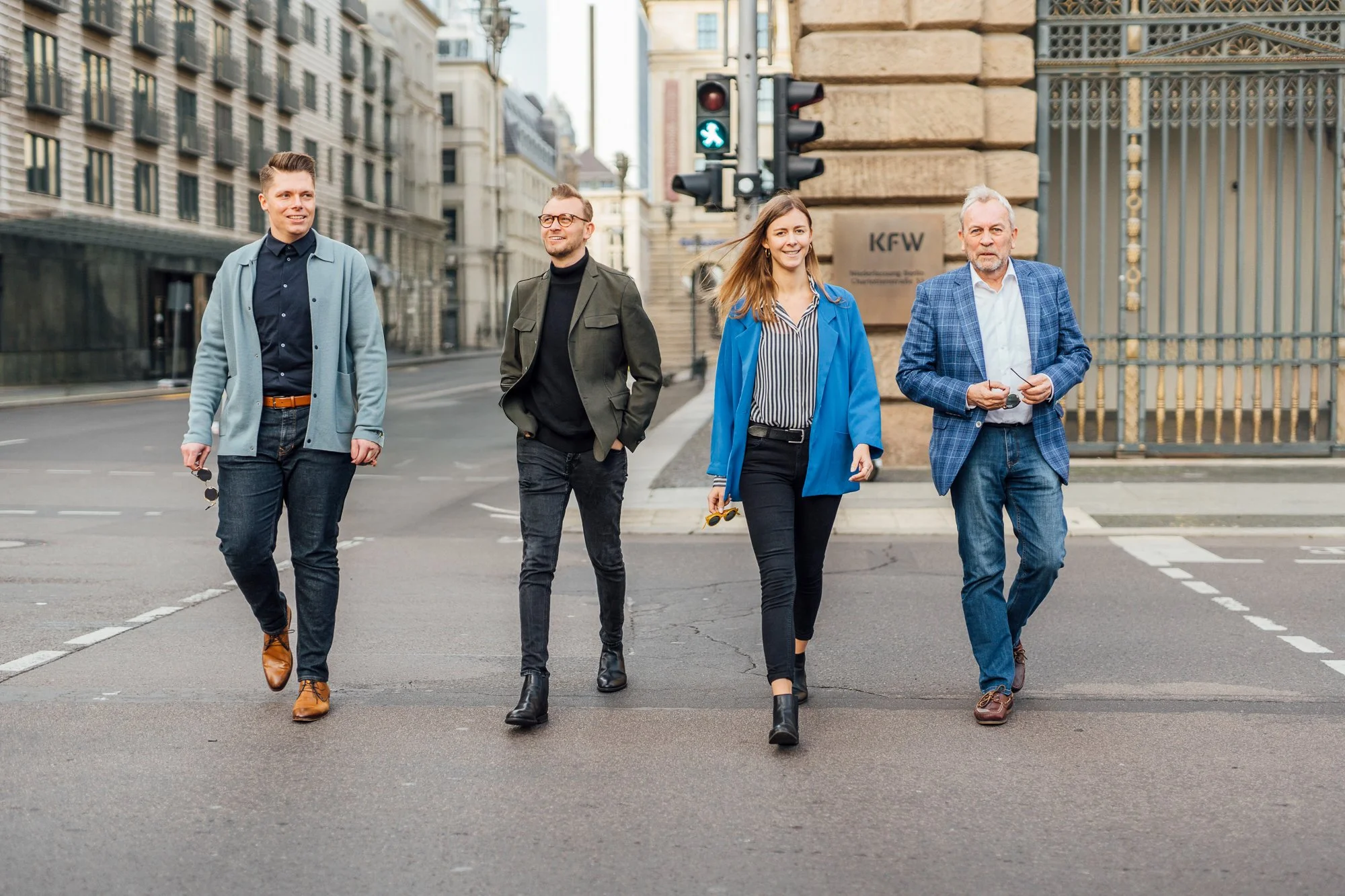 Vier Personen gehen durch eine Kreuzung in einer Stadt, mit Hochhäusern und einem großen Gittertor im Hintergrund, an einer Ampel, die grün zeigt.