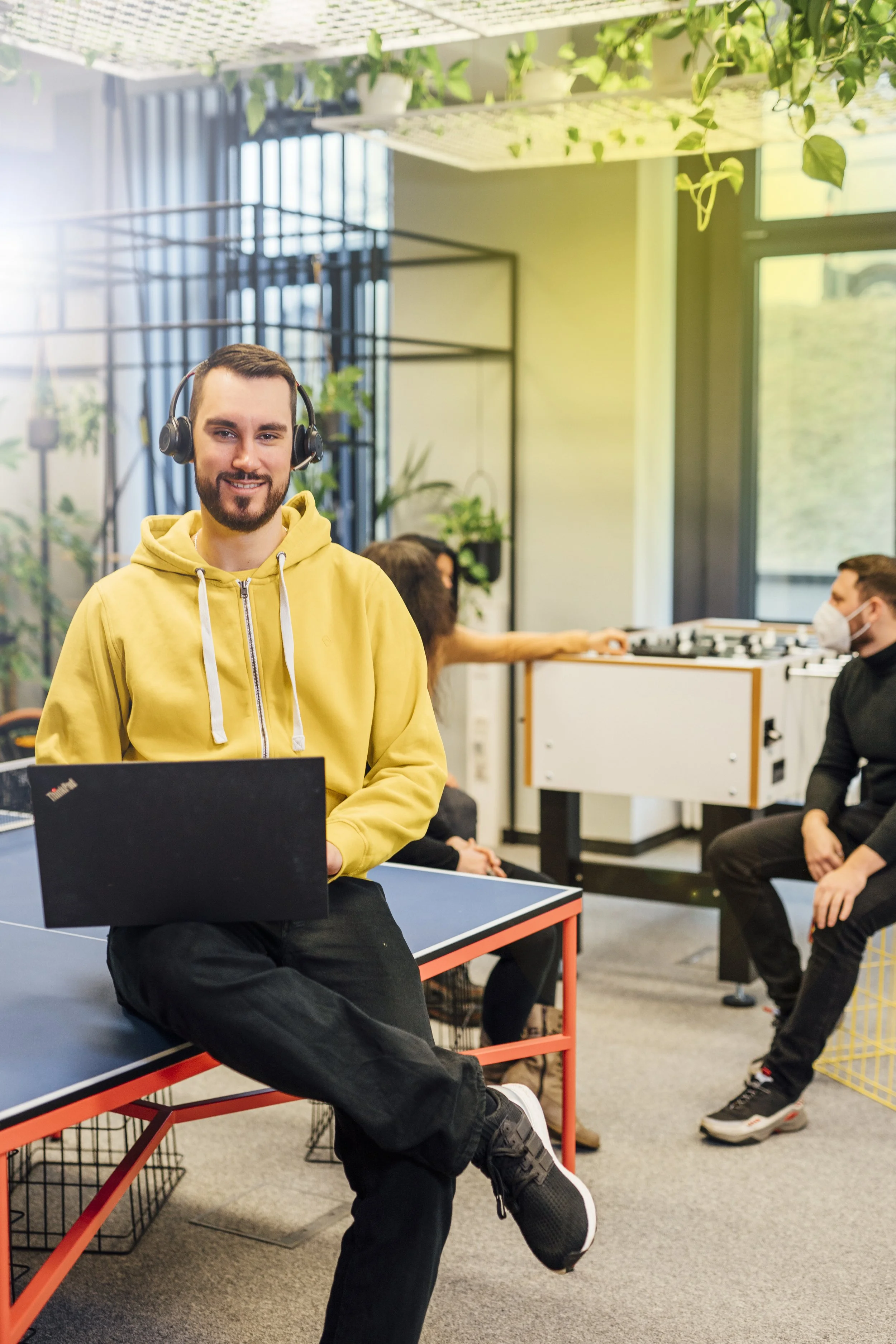 Junge Mann mit Headset, gelber Hoodie, sitzt auf Tisch in Gemeinschaftsbüro, im Hintergrund Familie beim Tischfußballspiel, andere Personen, ein Mann mit Maske, unterhalten sich.