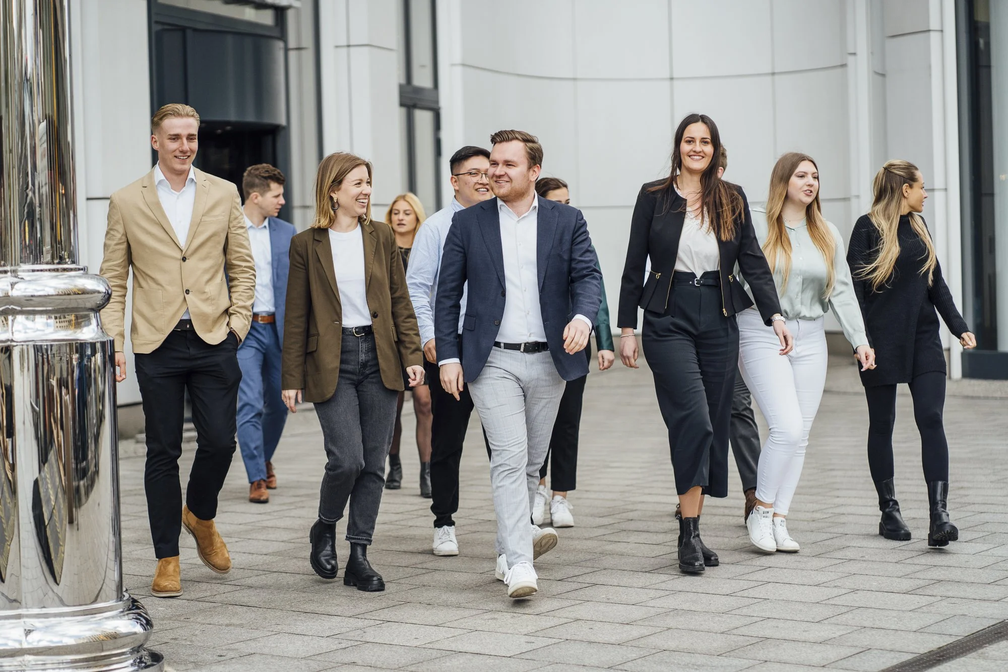 Eine Gruppe von jungen Erwachsenen geht lachend und in formeller Kleidung auf einer modernen Stadtstraße.