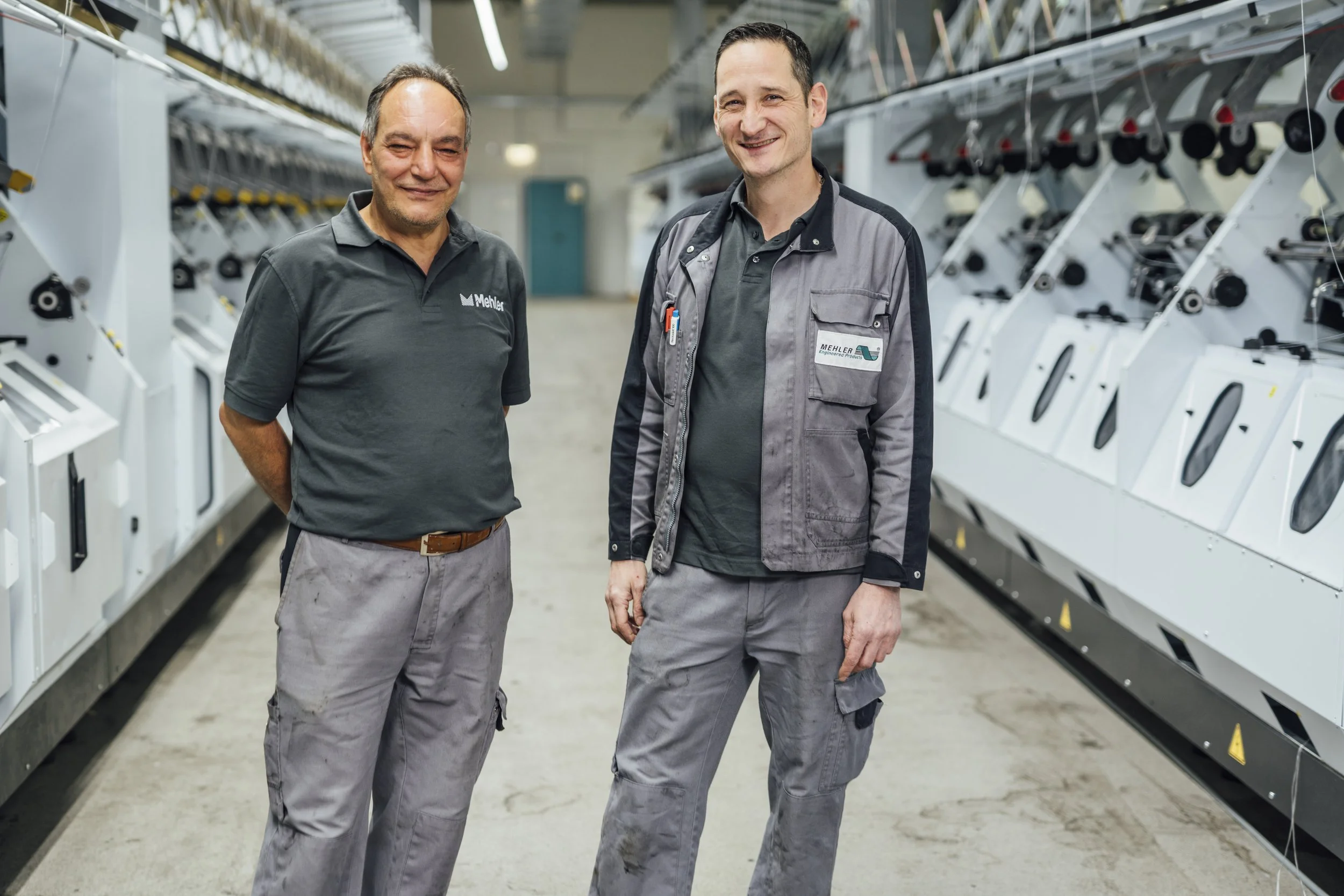 Zwei männliche Mitarbeiter stehen in einer Fabrikhalle neben industriellen Maschinen und lachen in die Kamera.