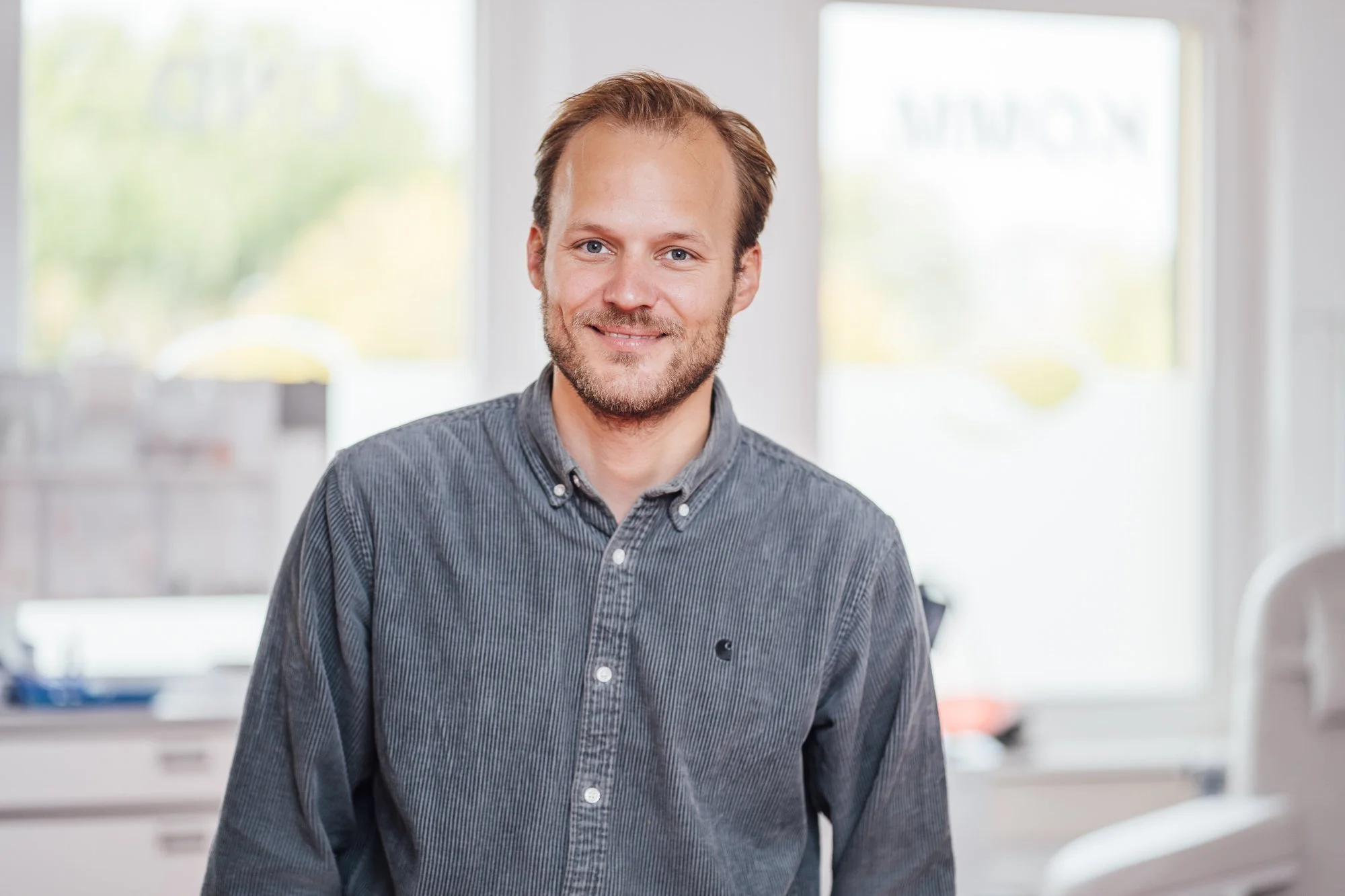 Ein lächelnder Mann mit braunen Haaren und Bart, trägt ein graues Hemd, steht in einem Büro mit großen Fenstern im Hintergrund.
