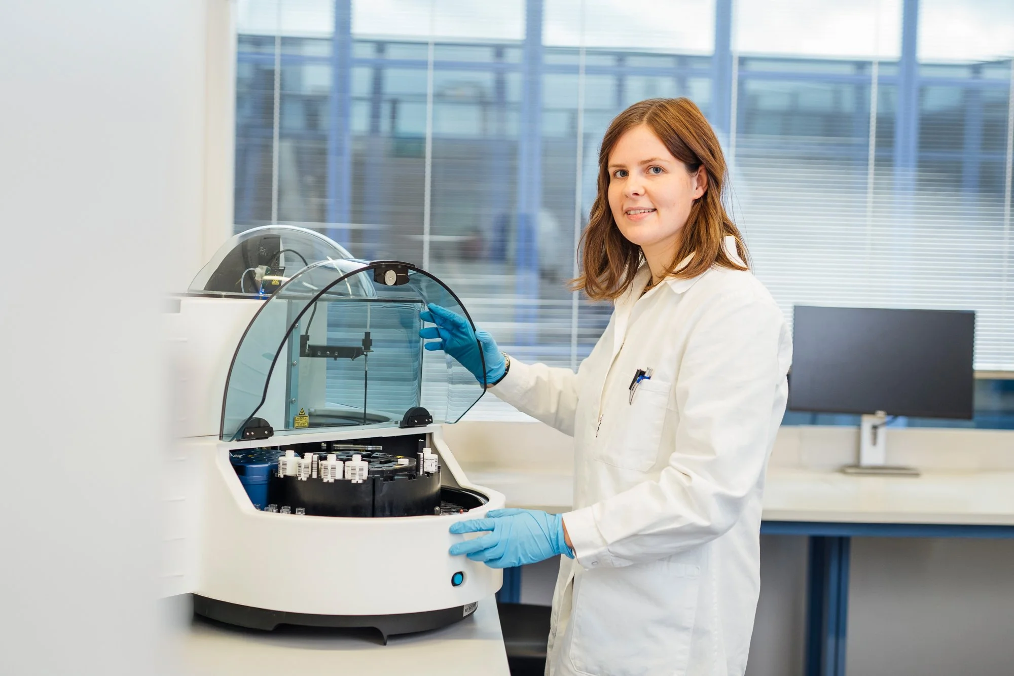 Eine Wissenschaftlerin in weißen Kittel und blaue Handschuhe bedient eine wissenschaftliche Maschine in einem Labor mit großen Fenstern im Hintergrund.