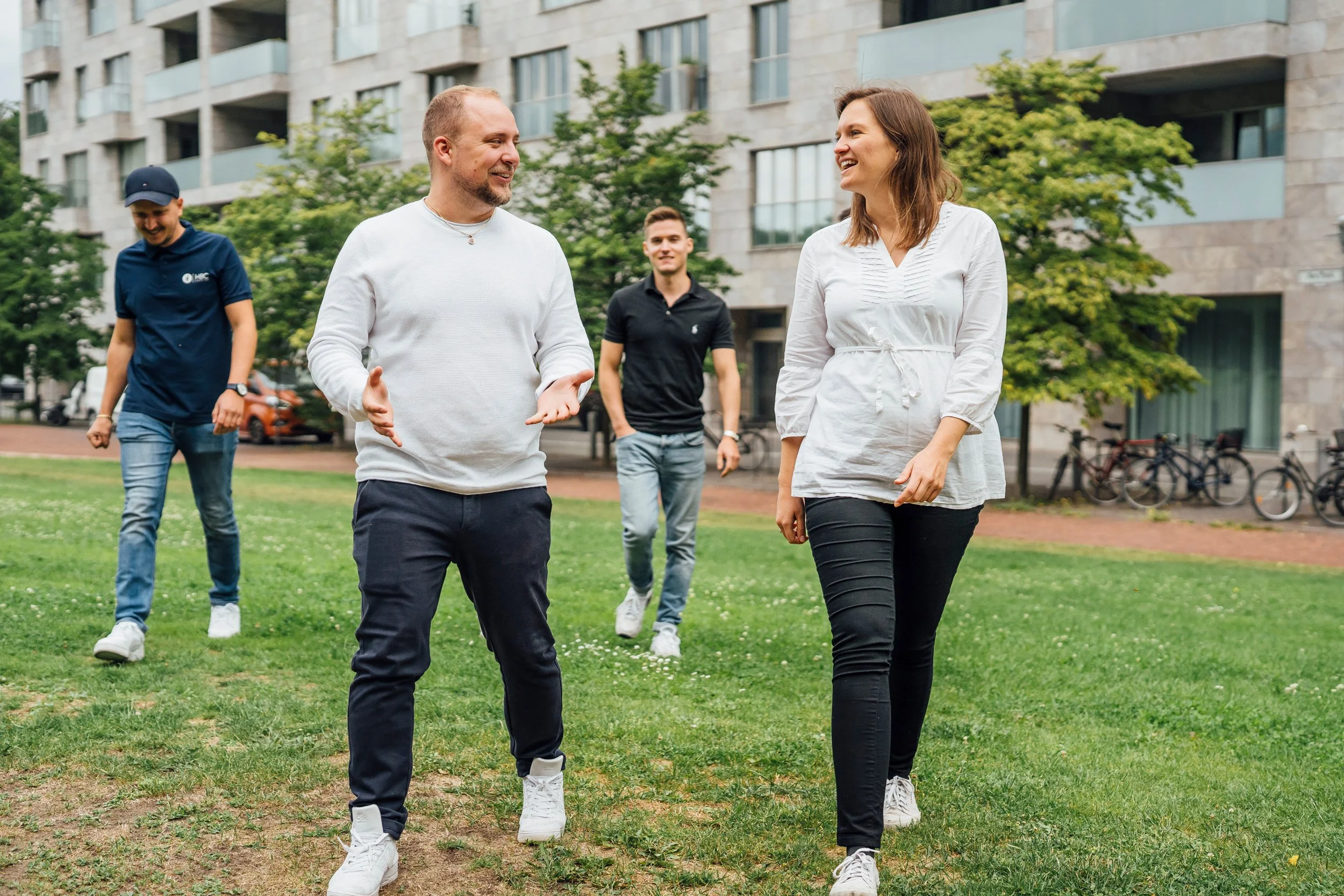 Fünf junge Menschen spazieren lachend durch einen Park in einer urbanen Umgebung mit modernen Gebäuden im Hintergrund.