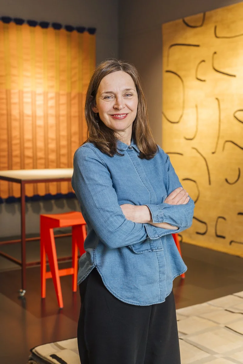 Eine lachende Frau mit braunen Haaren, die eine blaue Jeansbluse trägt, steht mit verschränkten Armen in einem Raum mit bunten Wanddekorationen.