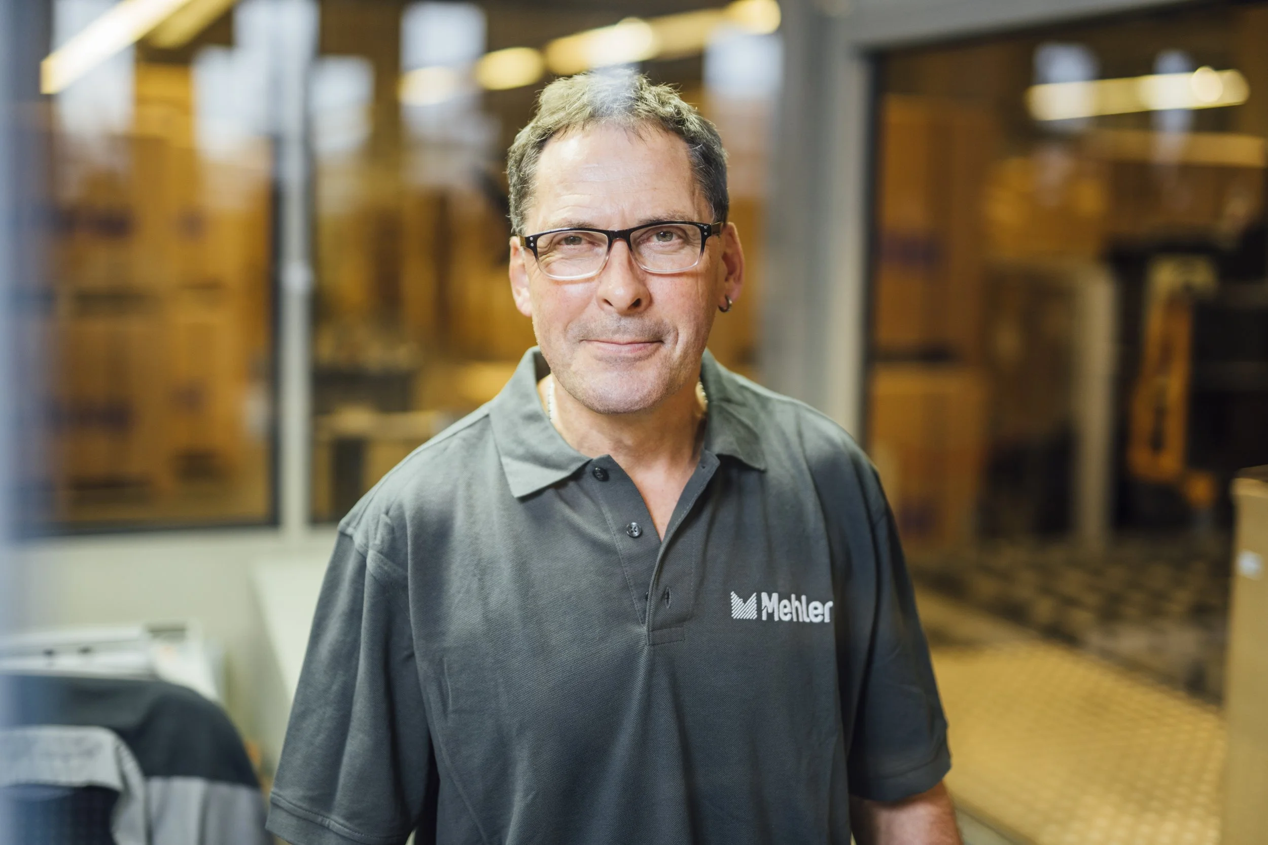 Ein Mann trägt ein dunkles Polo-Shirt mit dem Logo 'Mehler' und steht in einem Raum mit Holzwänden und Lagerregalen im Hintergrund.