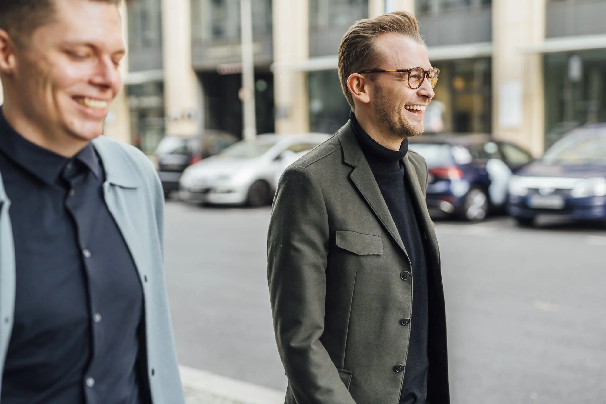 Zwei lachende Männer gehen auf einer Stadtstraße spazieren, im Hintergrund geparkte Autos und moderne Gebäude