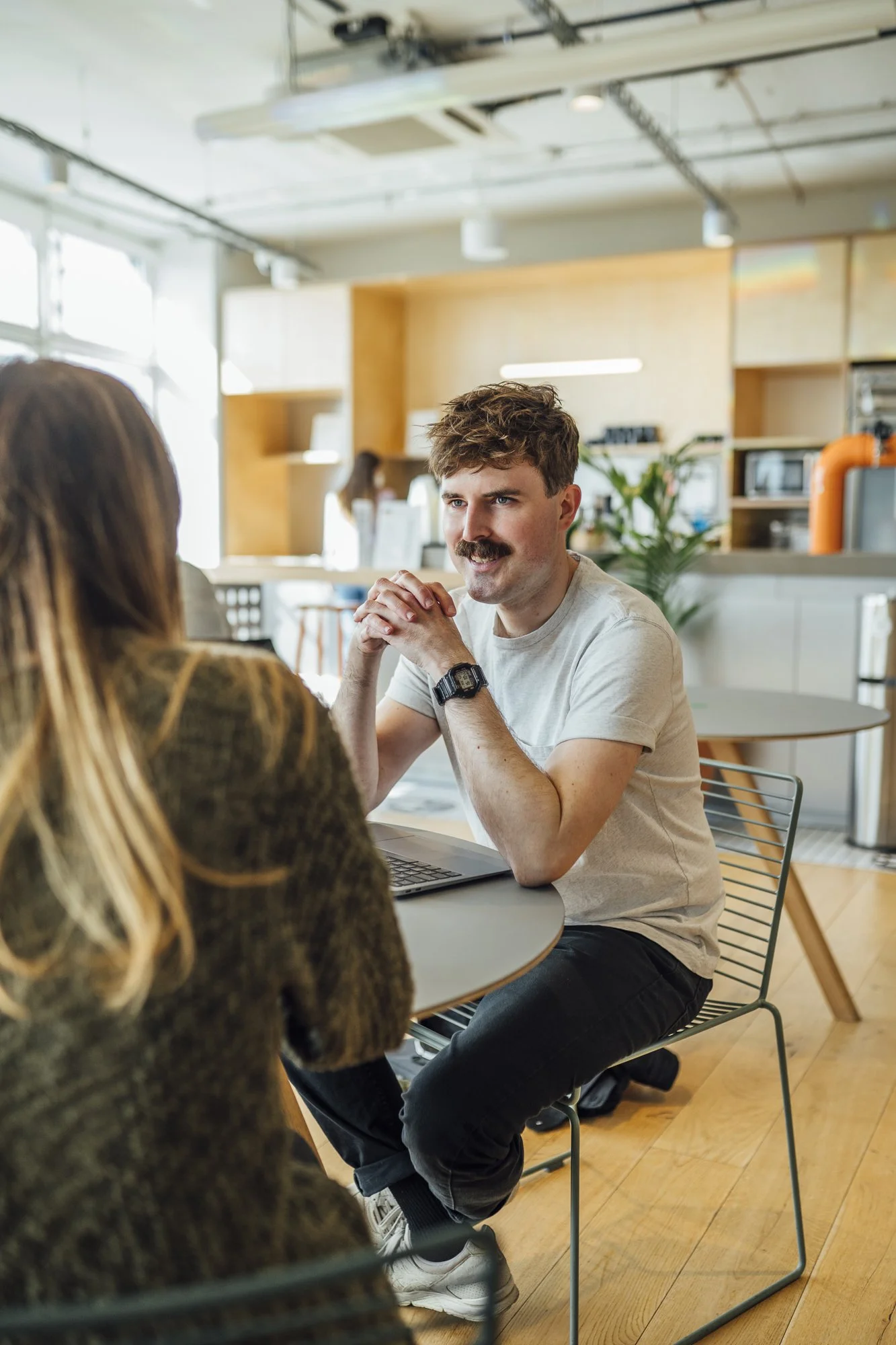 Zwei Menschen im Gespräch in einem modernen, hellen Büro, mit Laptop auf einem runden Tisch, im Hintergrund Küchenbereich mit Pflanzen.