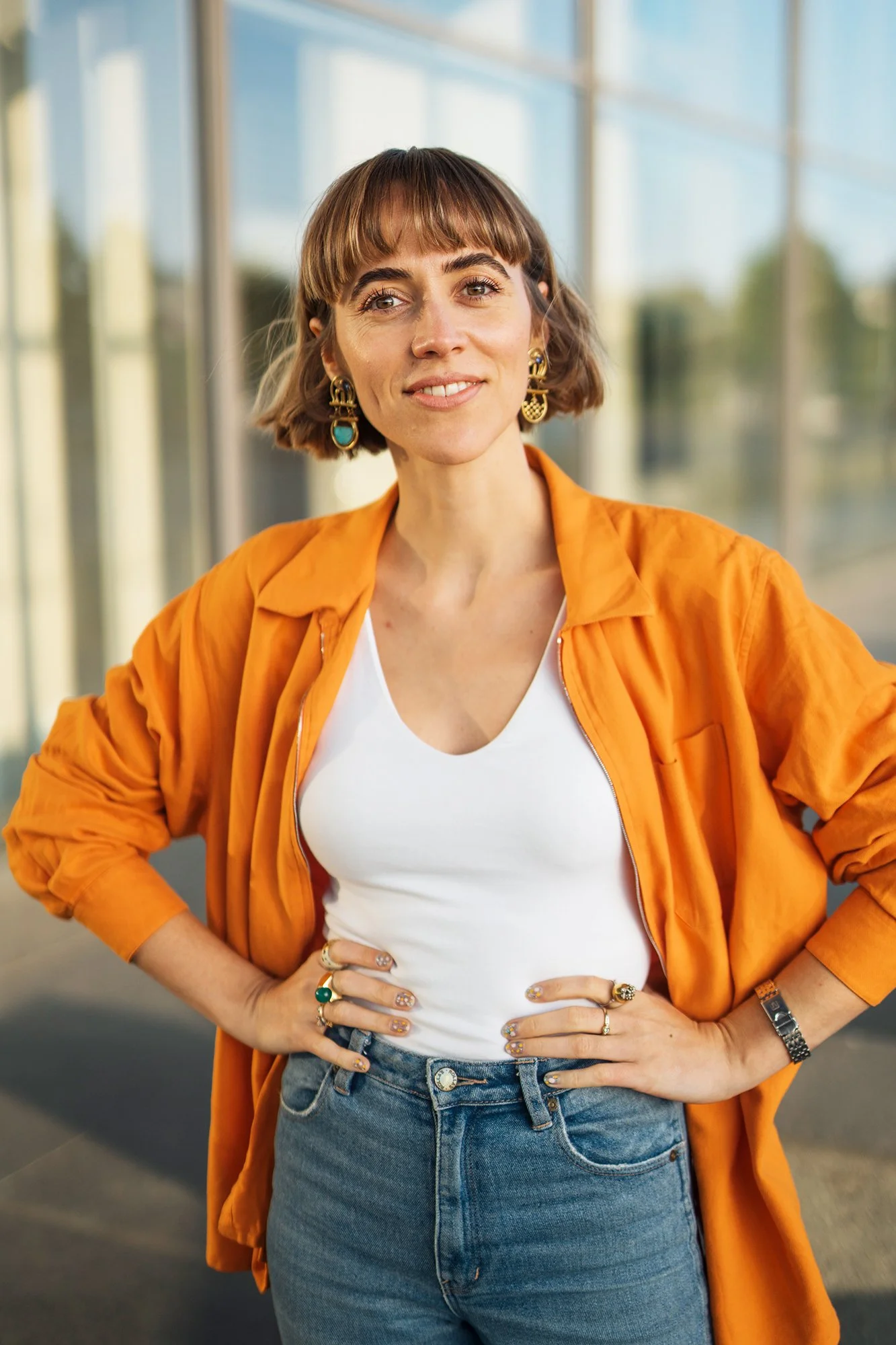 Eine junge Frau mit kurzem braunem Haar, lächelnd, trägt einen weißen T-Shirt, eine orange Jacke und Jeans, steht vor einem modernen Glasgebäude.