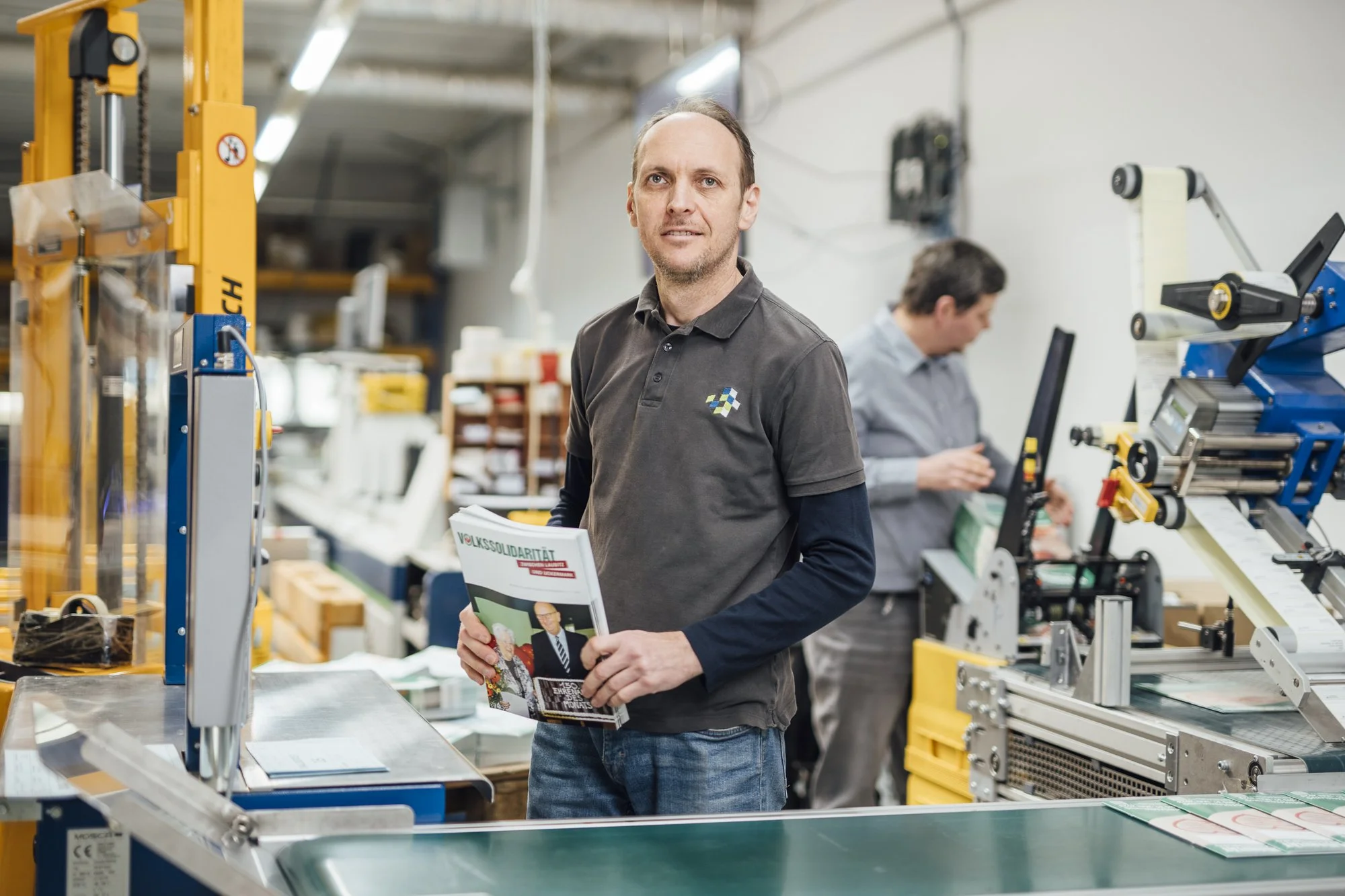 Zwei Männer arbeiten in einer Fabrikhalle bei Fertigungsmaschinen, eine Person hält eine Zeitschrift, beide tragen Hemden.