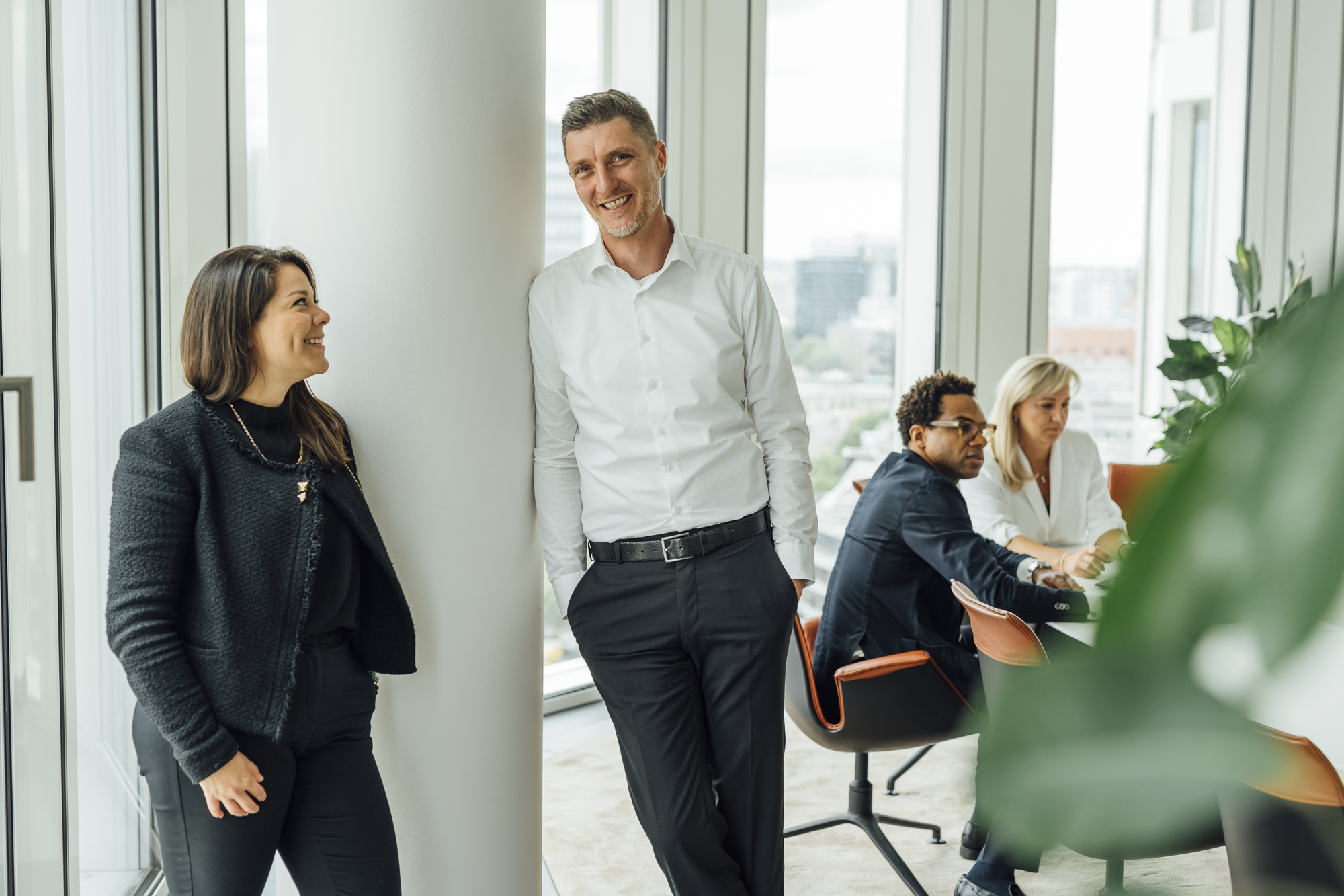 Vier Menschen in einem modernen Büro; zwei stehen und lachen, während zwei an Tischen arbeiten.