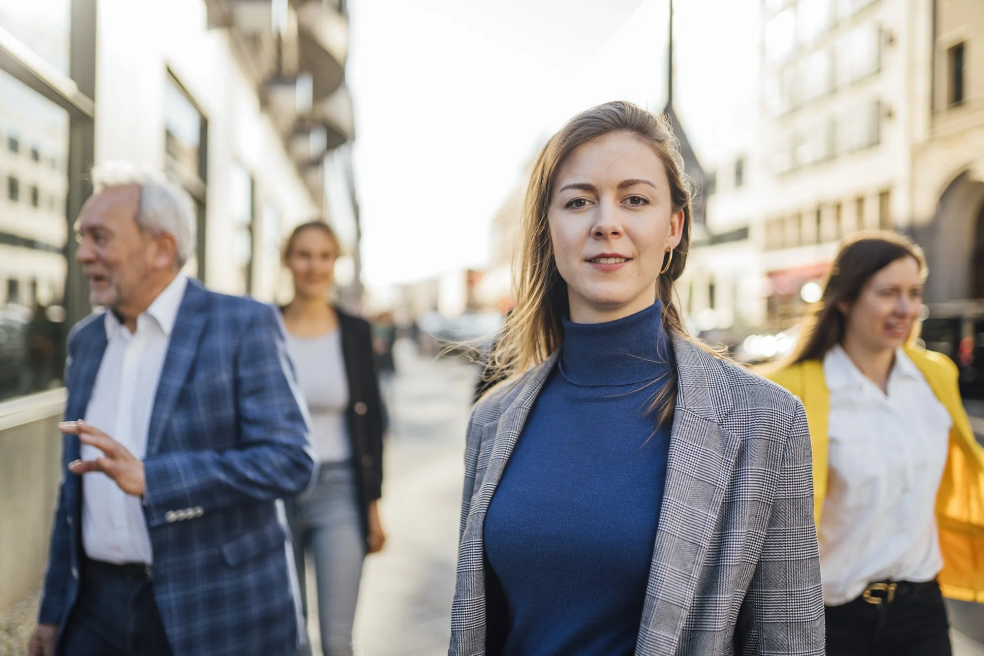 Vier Menschen gehen auf einer Stadtstraße, im Vordergrund eine junge Frau mit braunem Haar, blauer Rollkragen, grauem Blazer, lächelnd.