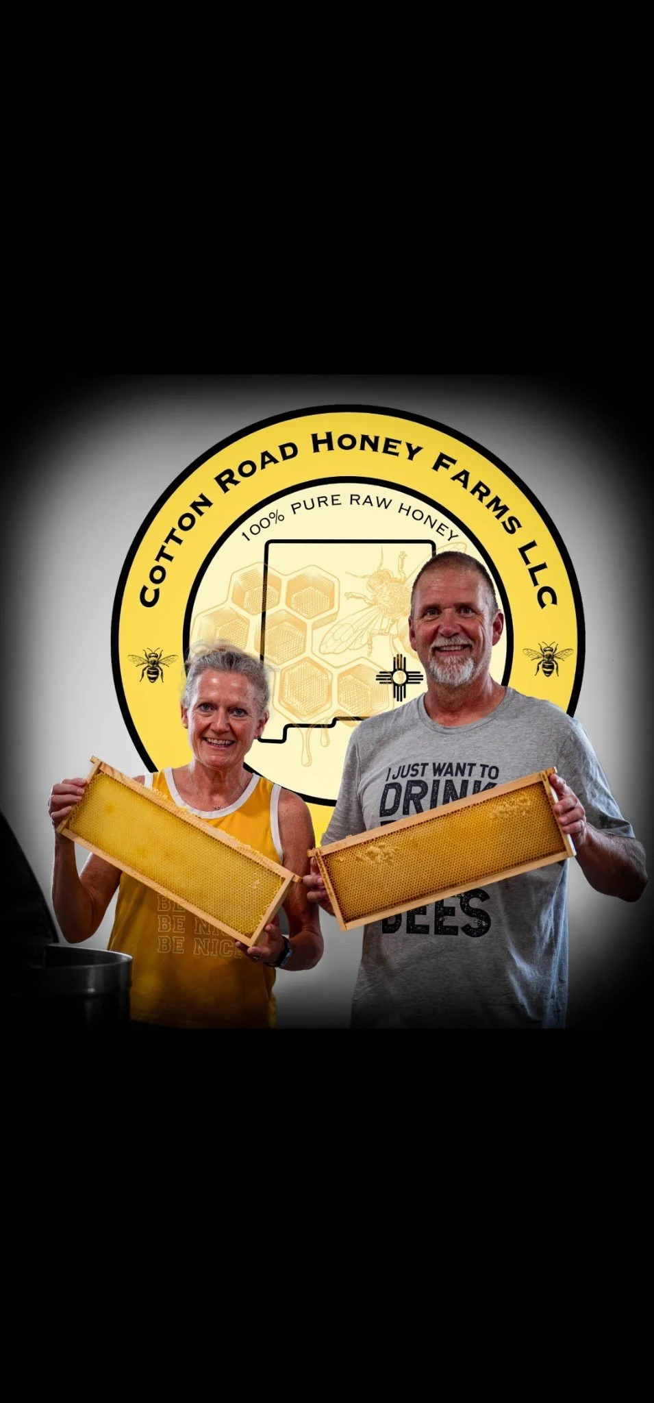 A man and woman holding honeycomb frames, standing in front of a sign for Cotton Road Honey Farms LLC that features a bee and honeycomb design. The woman is wearing a yellow shirt, and the man is wearing a gray shirt with text. Both are smiling.