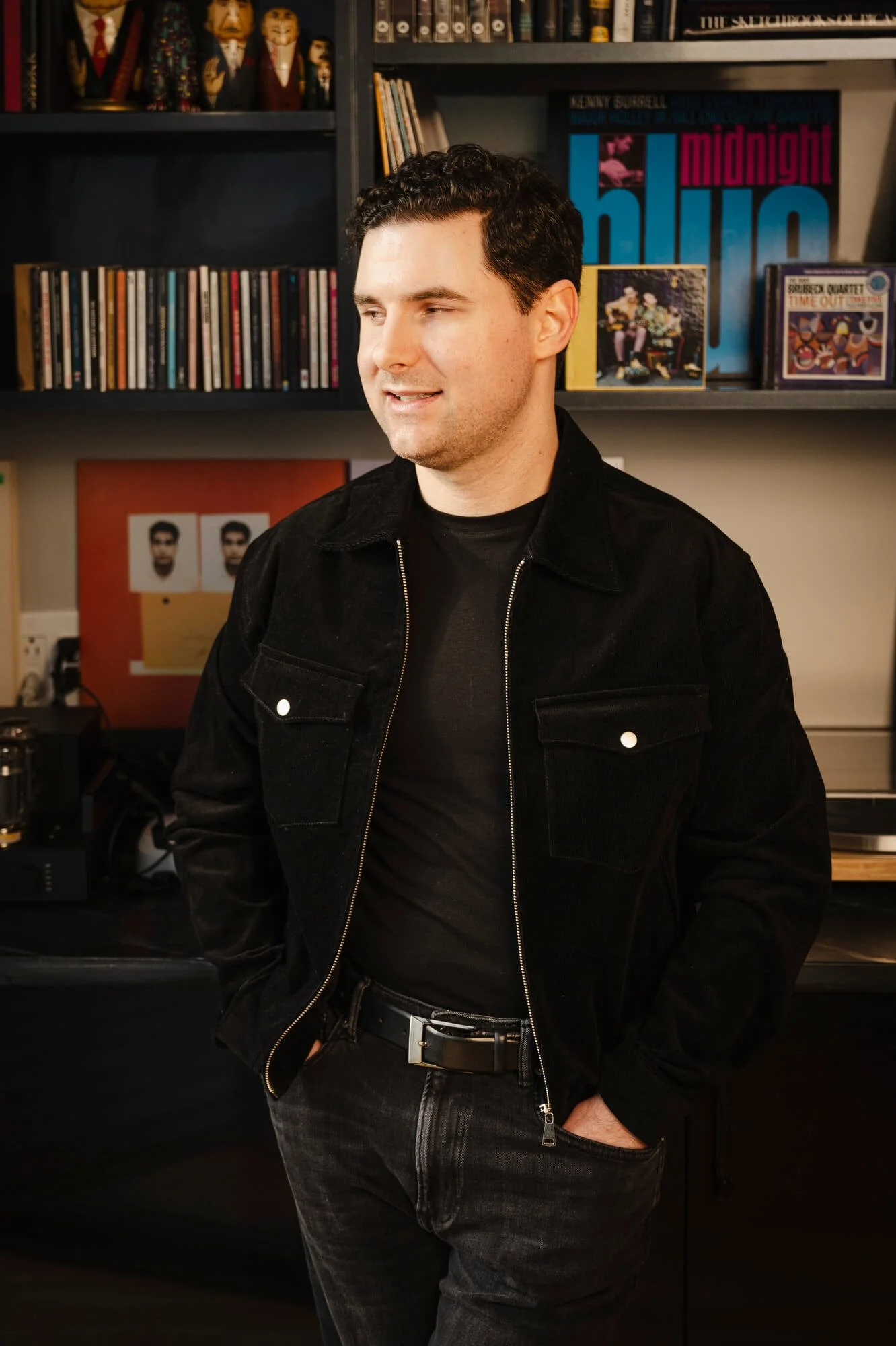 A young man with dark curly hair wearing a black jacket and black shirt, standing with his hands in his pockets in a room with shelves filled with books and vinyl records.