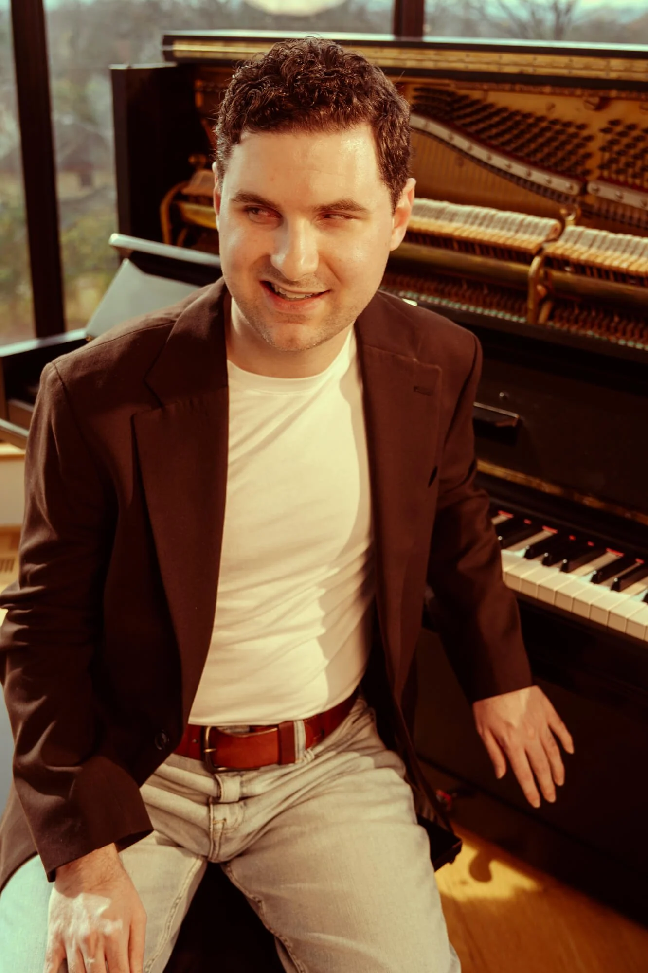 A man sitting by a piano in a room with large windows, wearing a brown blazer, white t-shirt, and beige pants.-- Elliott McClain- Elliottmclain.com
