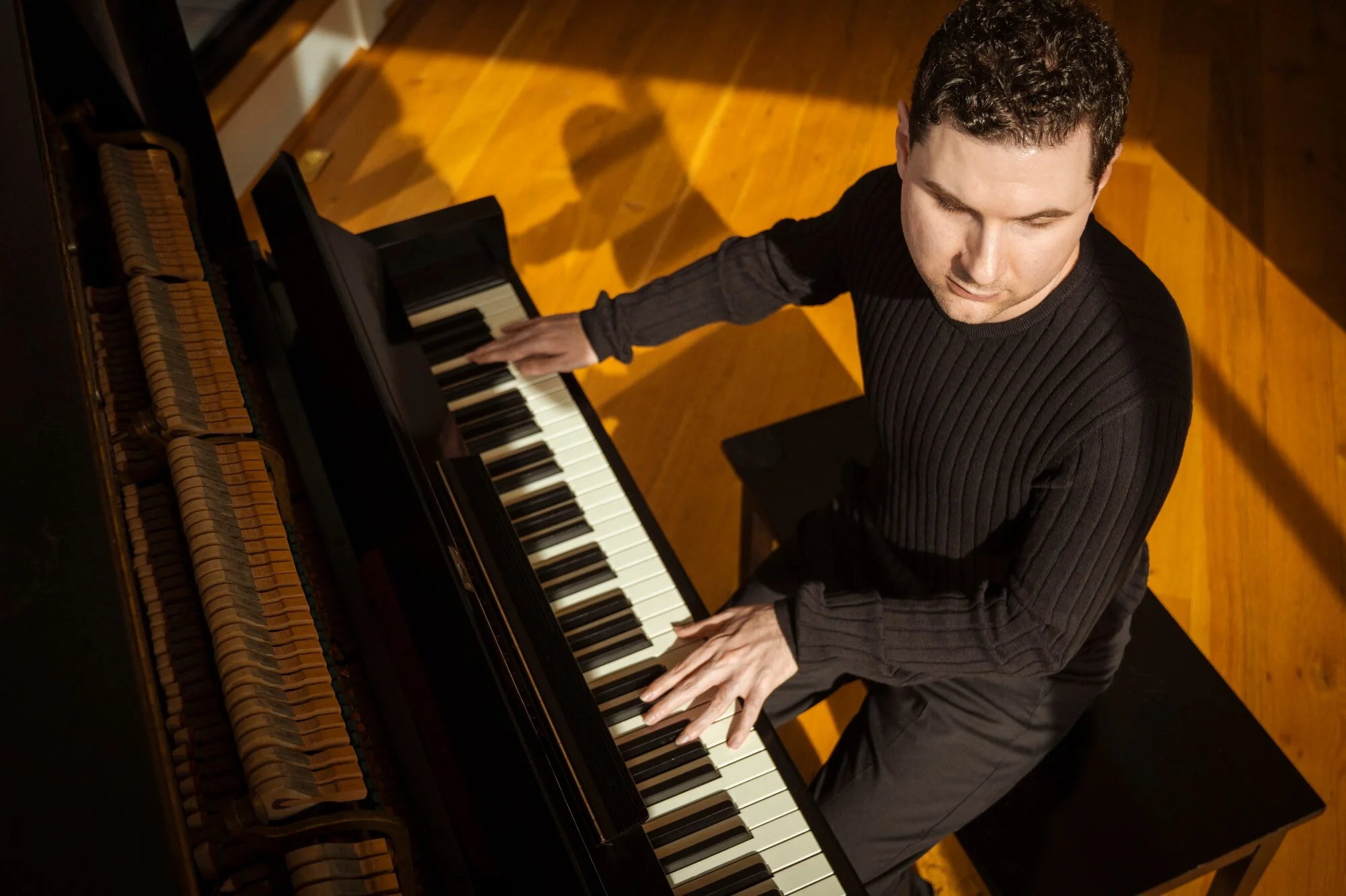 A young man with short curly hair playing a black grand piano, viewed from above in a room with wooden floors.
