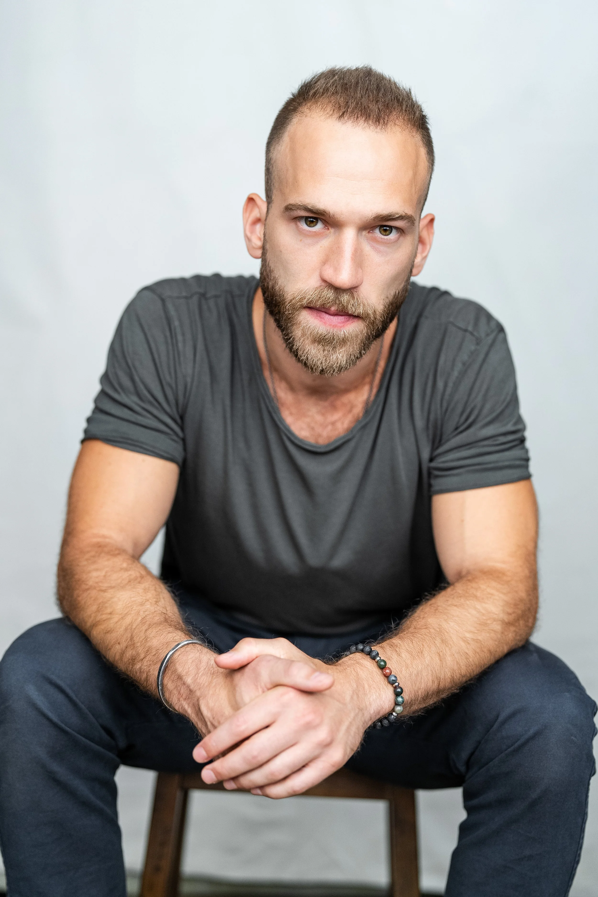 A man with short hair and a beard, wearing a gray t-shirt, sitting on a wooden chair against a plain white wall, with his hands clasped together in front of him.