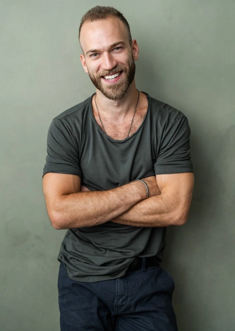 Smiling man with short hair and beard, wearing a dark gray t-shirt and dark pants, standing against a plain background with arms crossed.