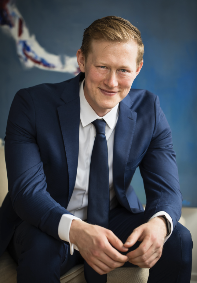 A young man in a navy suit with a white shirt and blue tie, sitting and smiling indoors.