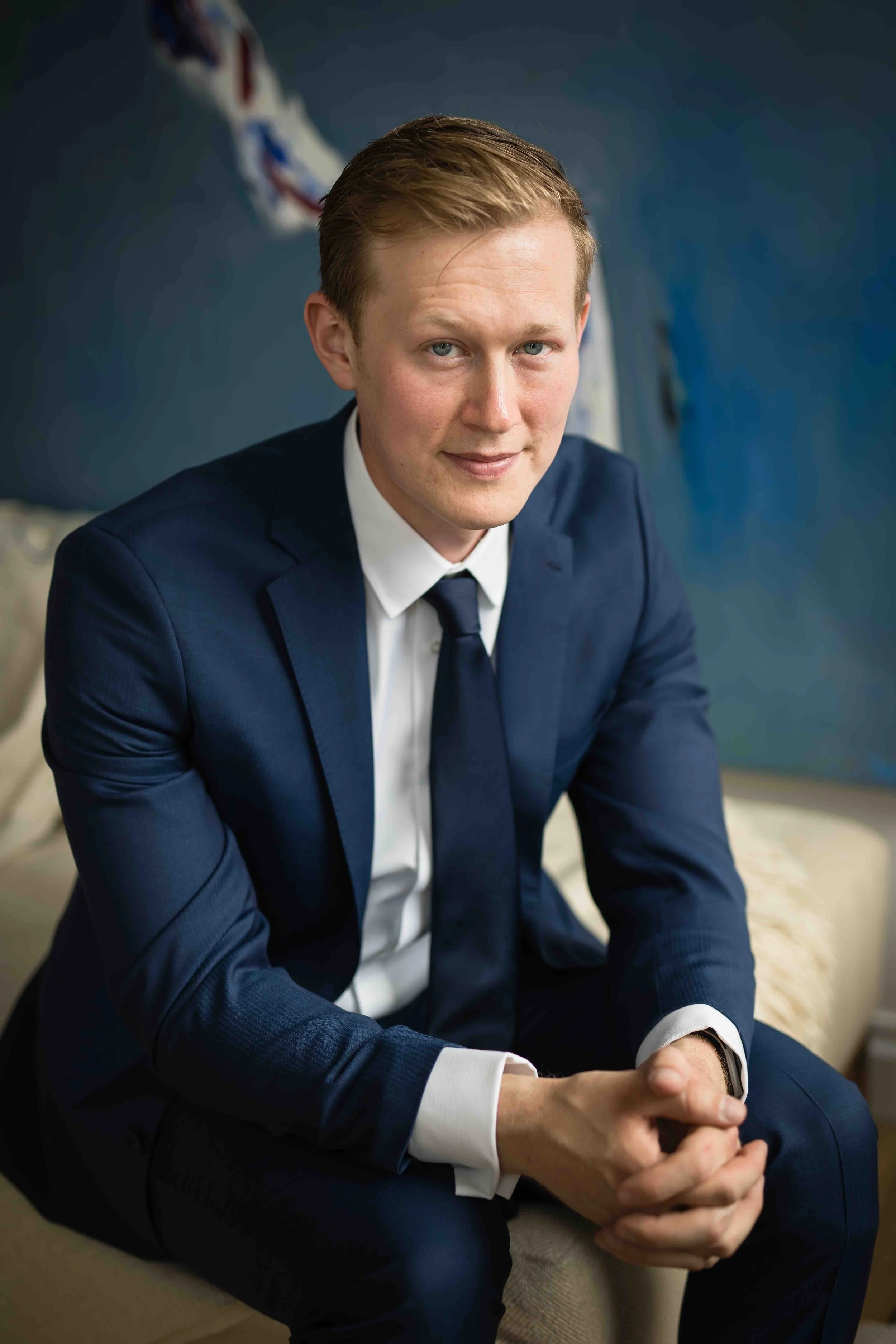 A man in a dark blue suit with a white shirt and dark blue tie, sitting on a beige couch, clasping his hands together, smiling slightly, with a blue wall in the background.