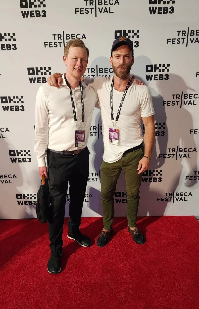 Two men standing on a red carpet at Tribeca Film Festival, posing for a photo with their arms around each other, in front of a step-and-repeat backdrop with the event's logo.