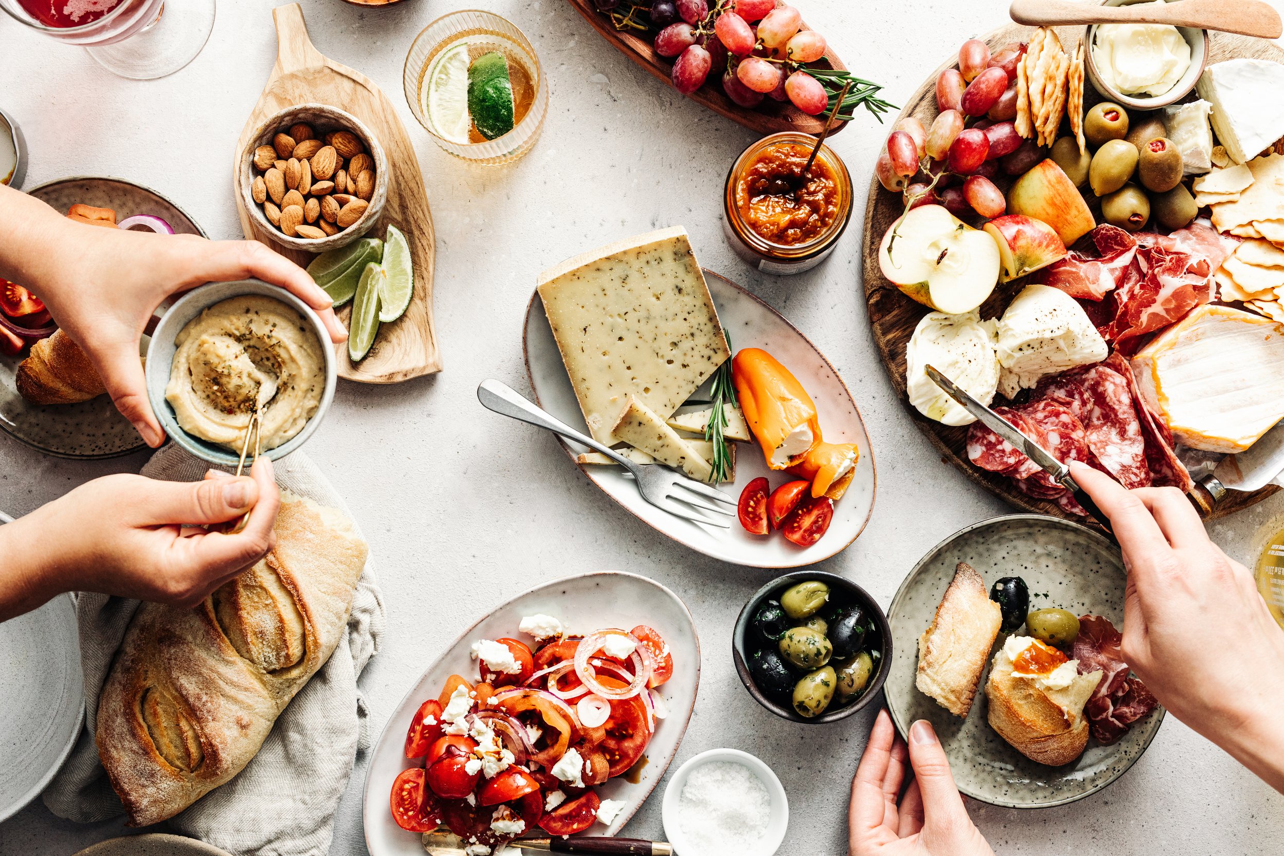 A cheese platter with various cheeses, grapes, apple slices, almonds, and crackers. Someone is spreading hummus on a piece of bread, with additional bread rolls and a plate of tomato and feta salad nearby. There are also glasses of drinks, olives, and someone slicing cheese.