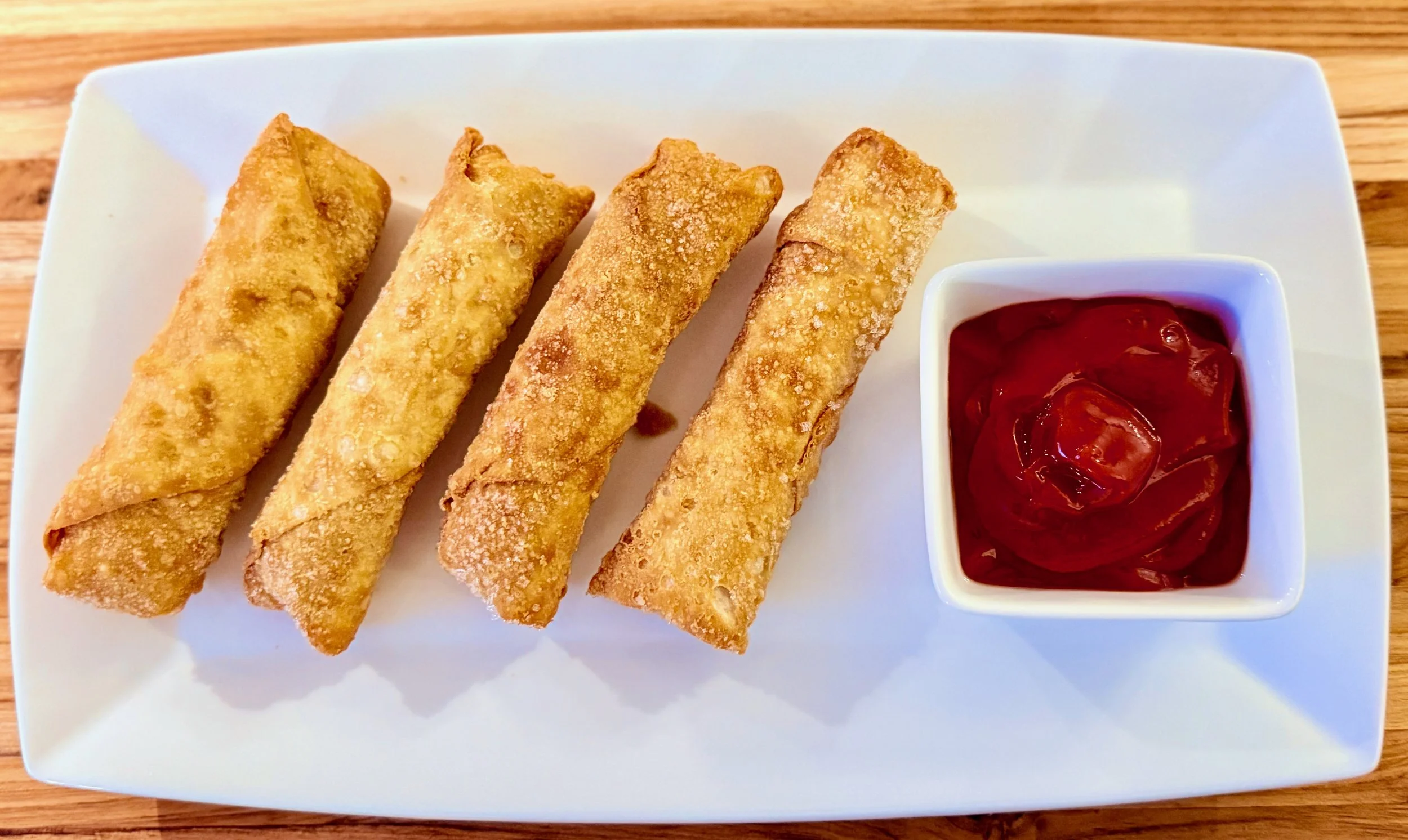 Four fried egg rolls on a white rectangular plate with a side of red dipping sauce in a small white bowl, all on a wooden table.