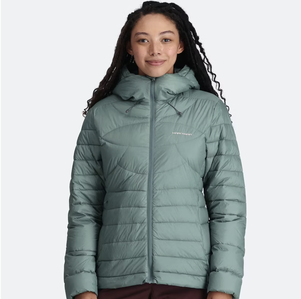 A woman with long, curly hair and a septum piercing wearing a teal puffer jacket with a hood.