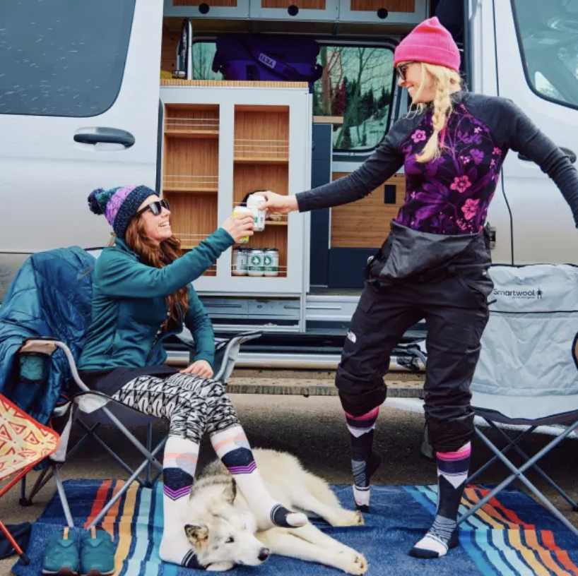 Two women in colorful winter clothing exchanging pills or supplements in front of a camper van, with one sitting on a camping chair and the other standing, while a dog lies on a blanket on the ground.