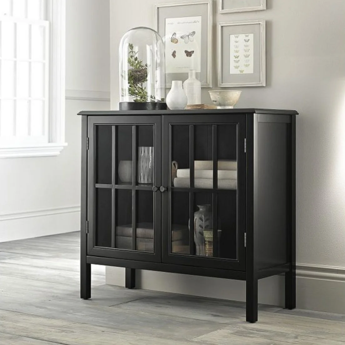 Black cabinet with glass doors displaying books and vases, decorated with white vases, a glass cloche with a plant, and framed butterfly art on a white wall.