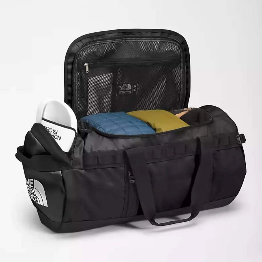 Black North Face duffel bag with its zipper open, showing packed clothing and a shoe in the side pocket on a white background.
