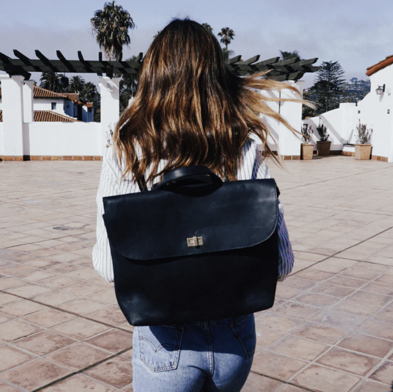 A woman with shoulder-length brown hair walking outdoors on a tiled patio, carrying a black backpack, with white buildings and palm trees in the background.