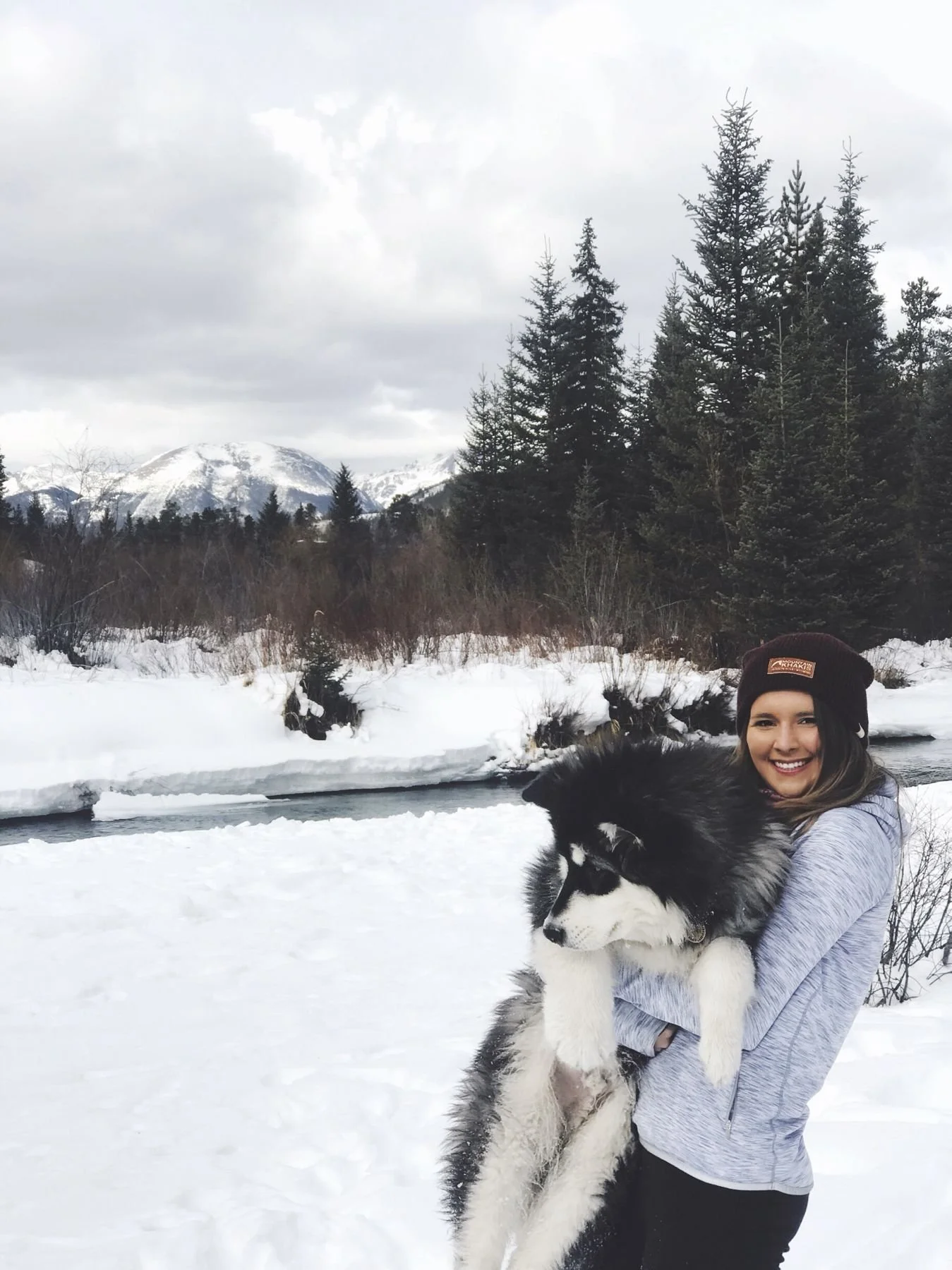 A smiling woman in a gray jacket and black beanie holding a large black and white Siberian Husky puppy in a snowy landscape with trees and mountains in the background.